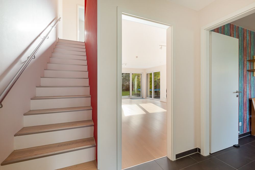 Flur mit Treppe, offenem Durchgang zum Wohnzimmer mit großen Fenstern und Zugang zu einem weiteren Raum.