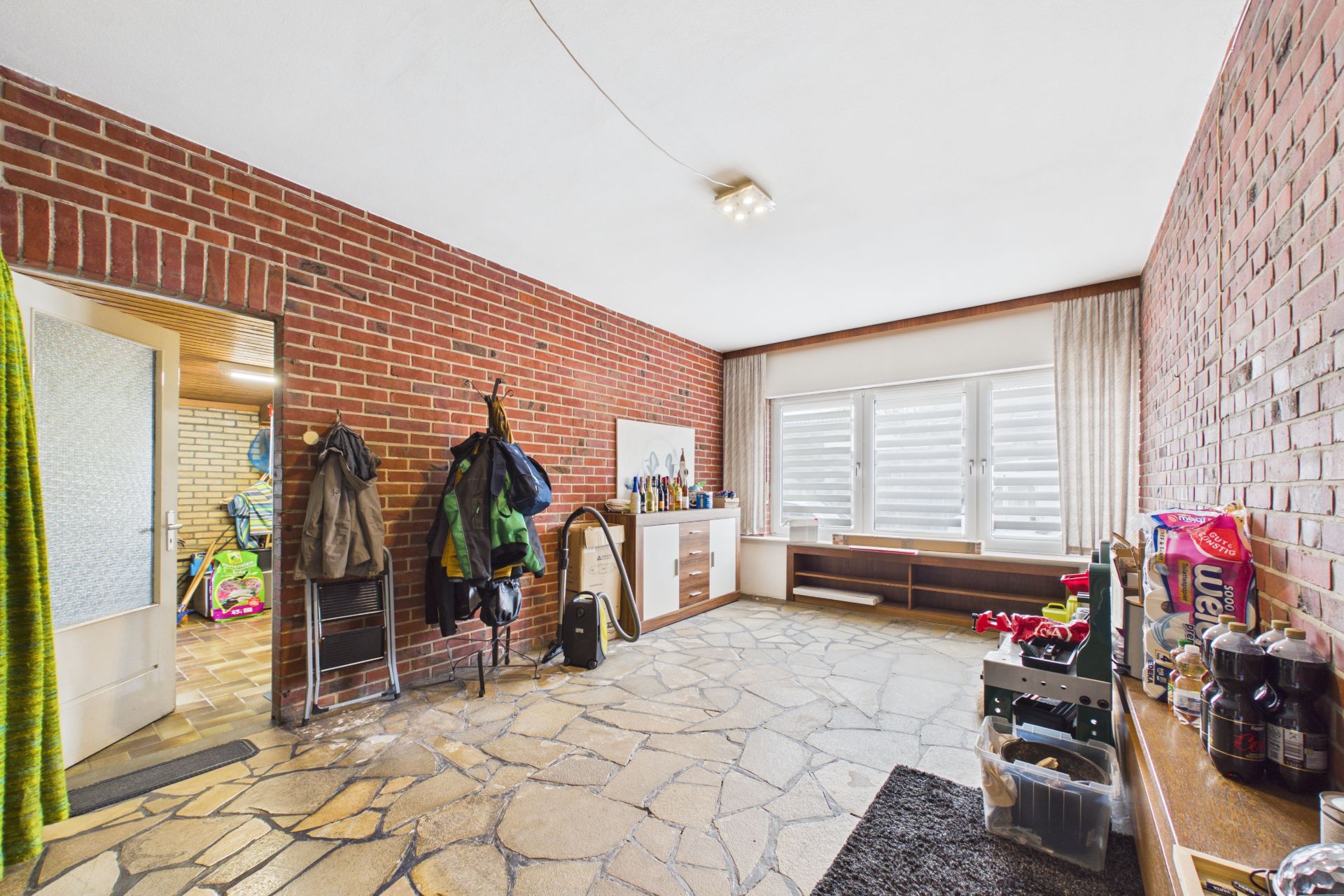Zimmer mit roten Backsteinwänden, Garderobe, Steinfussboden und großem Fenster mit weißen Vorhängen.