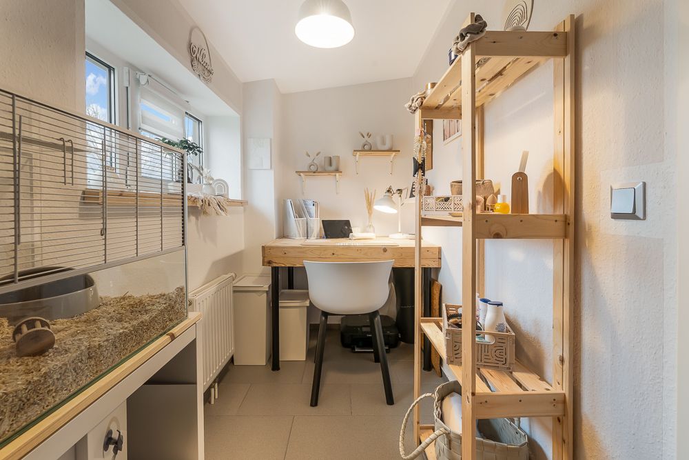 Kleines Arbeitszimmer mit Schreibtisch, Regal und großem Haustierkäfig am Fenster.