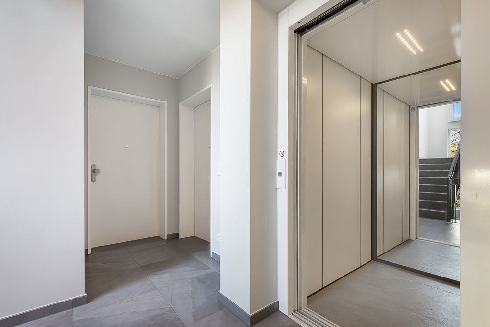 Heller Flur mit offenem Aufzug, geschlossener Tür und Treppe im Hintergrund.