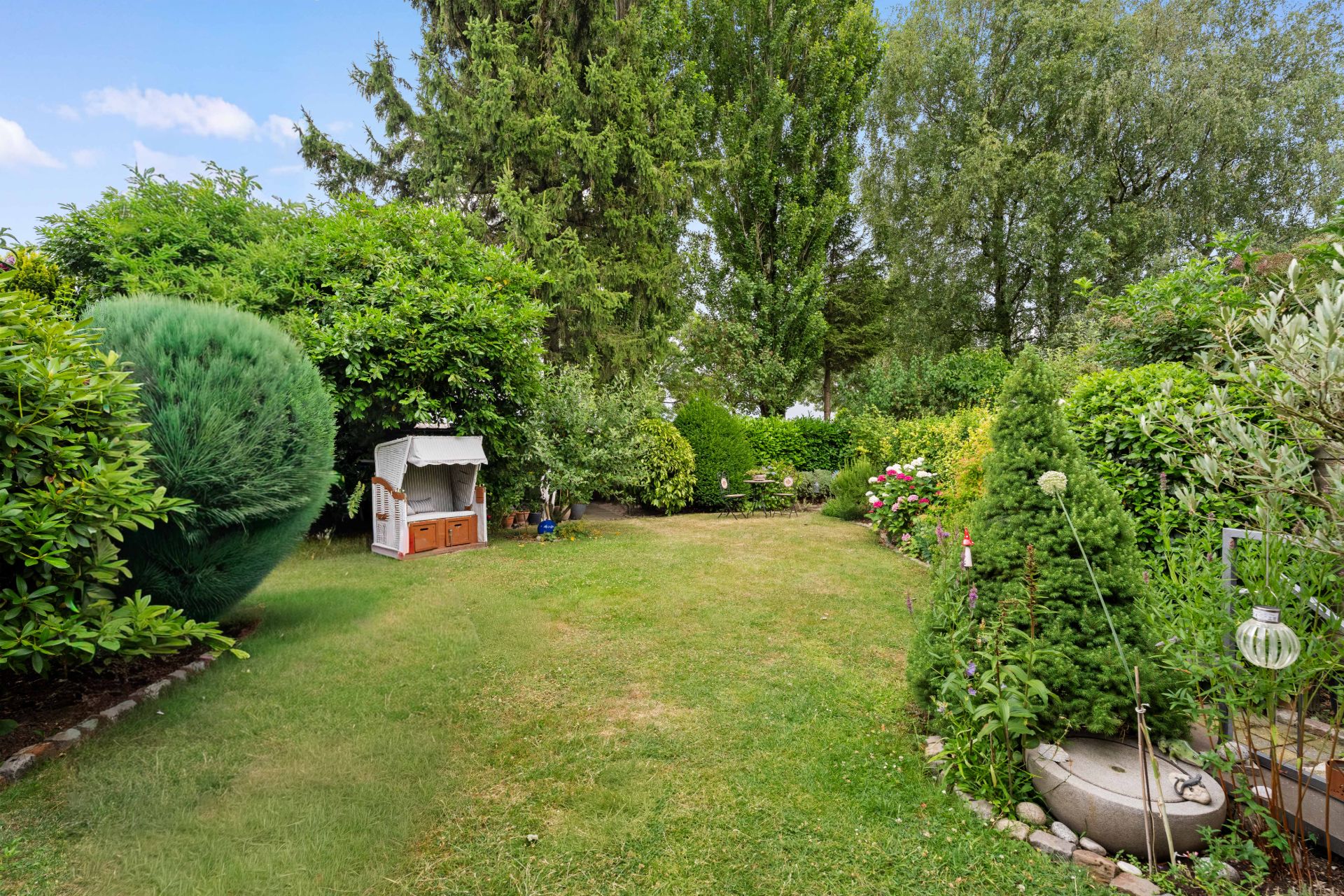 Großer Garten mit vielen Sträuchern, Bäumen und einem Strandkorb auf einer Rasenfläche.