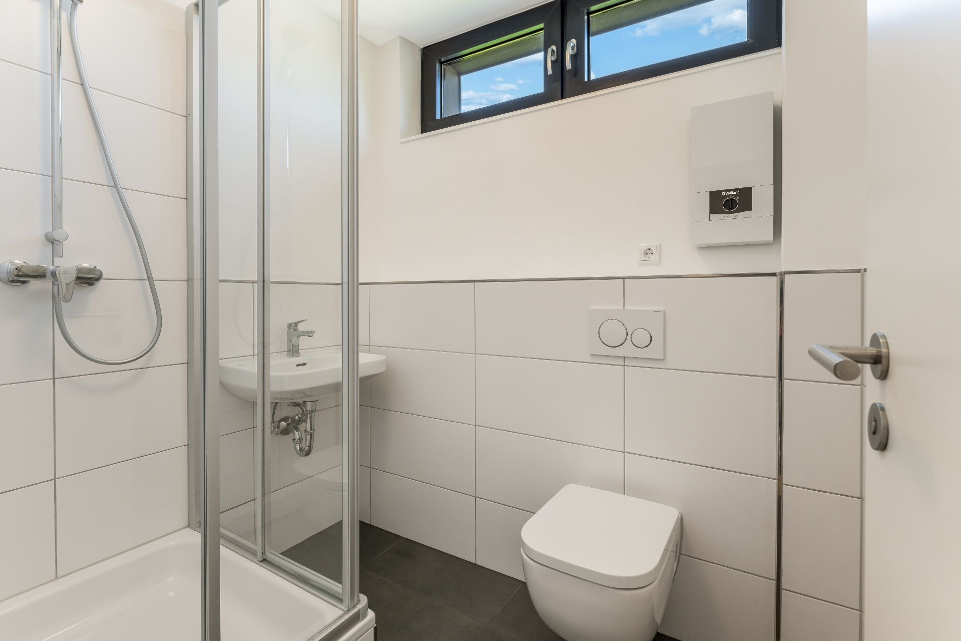 Modernes, helles Badezimmer mit Dusche, WC, kleinem Waschbecken und Fenster.