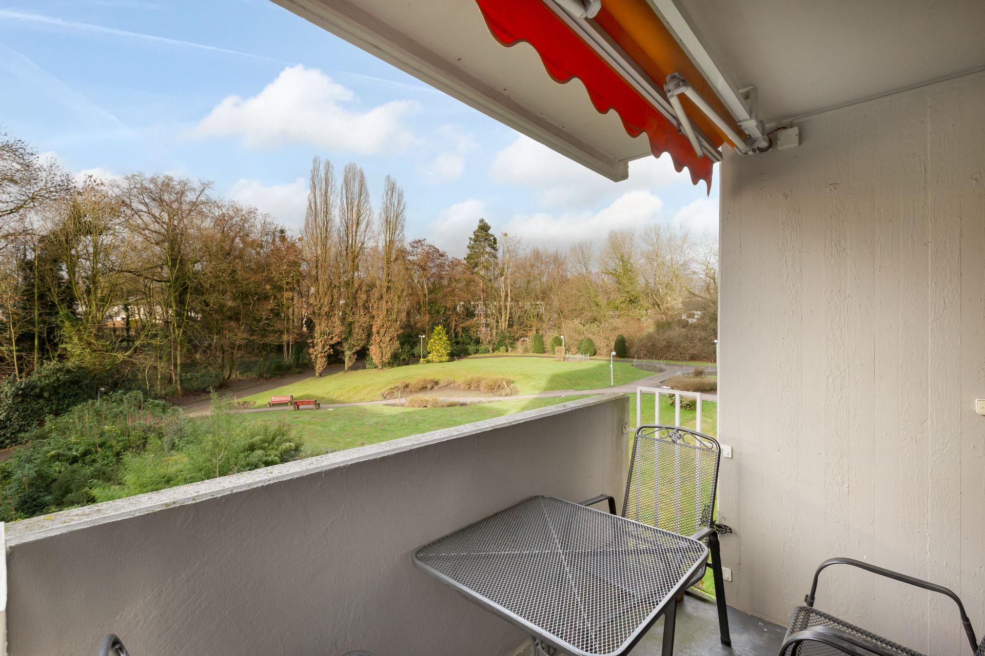 Balkon mit Tisch, zwei Stühlen und Markise, Blick auf eine grüne Parklandschaft.