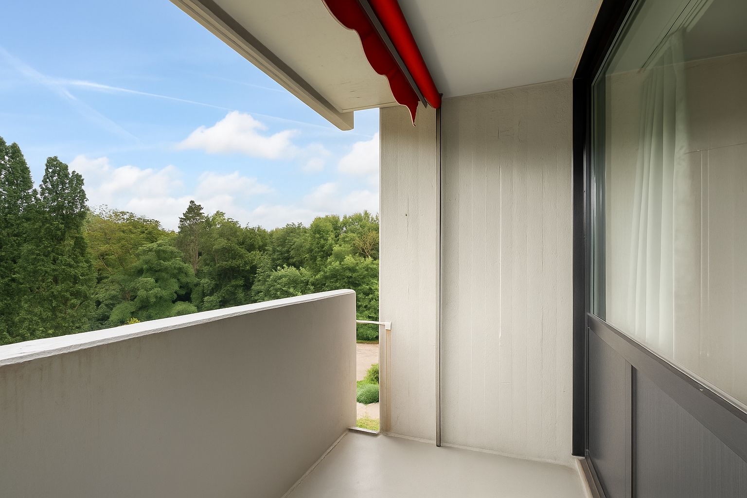 Balkon mit rotem Sonnenschutz und Blick auf grüne Bäume.