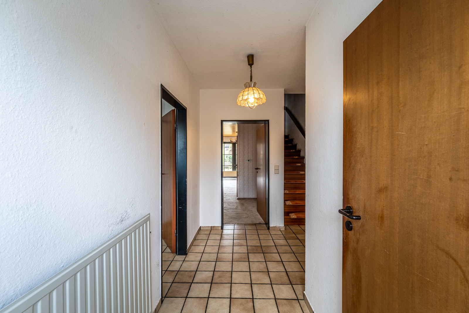 Flur mit Fliesenboden, Holztüren und Treppe, Lampenschirm an der Decke.
