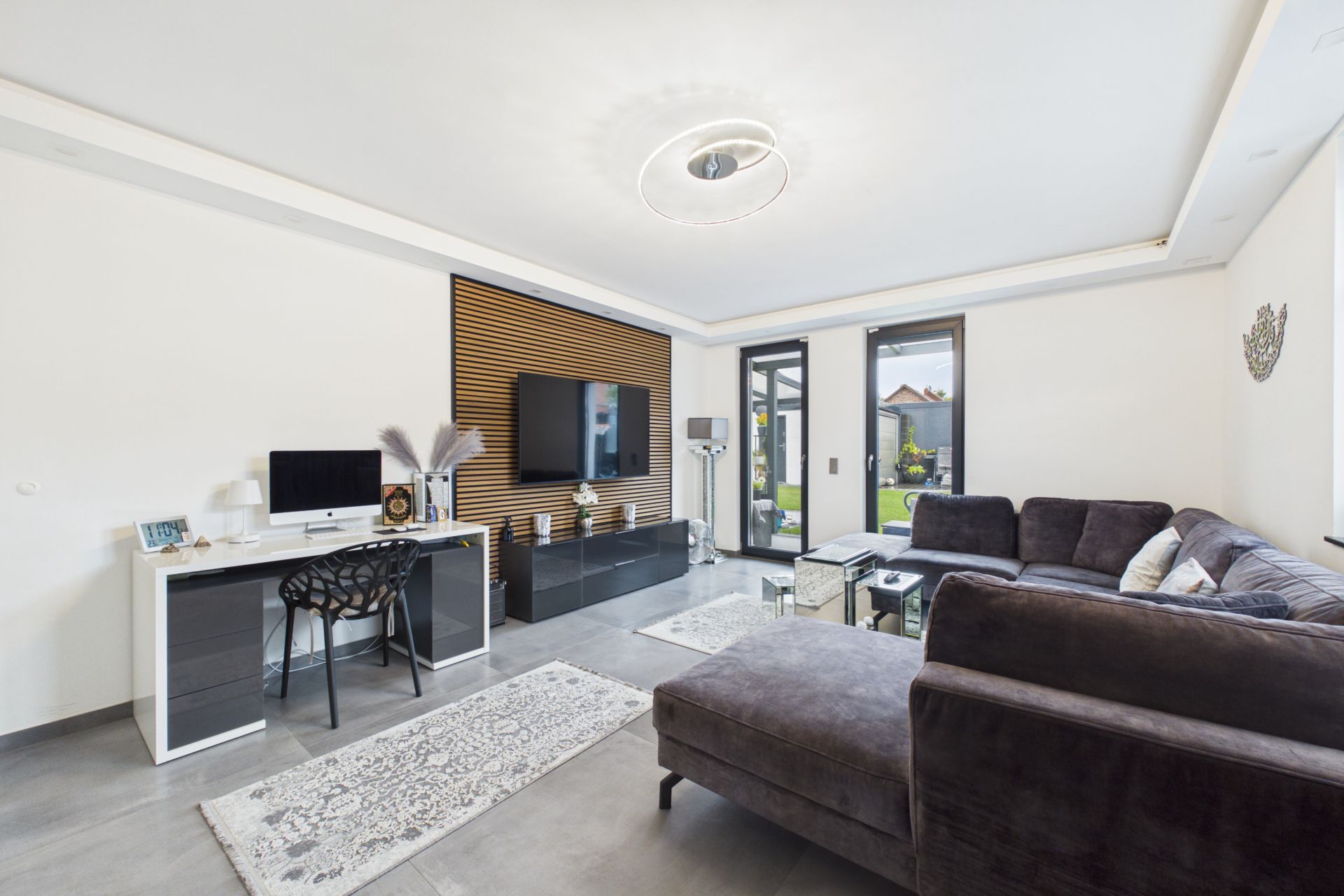 Modernes Wohnzimmer mit Ecksofa, Schreibtisch, Fernseher und großen Fenstern zum Garten.