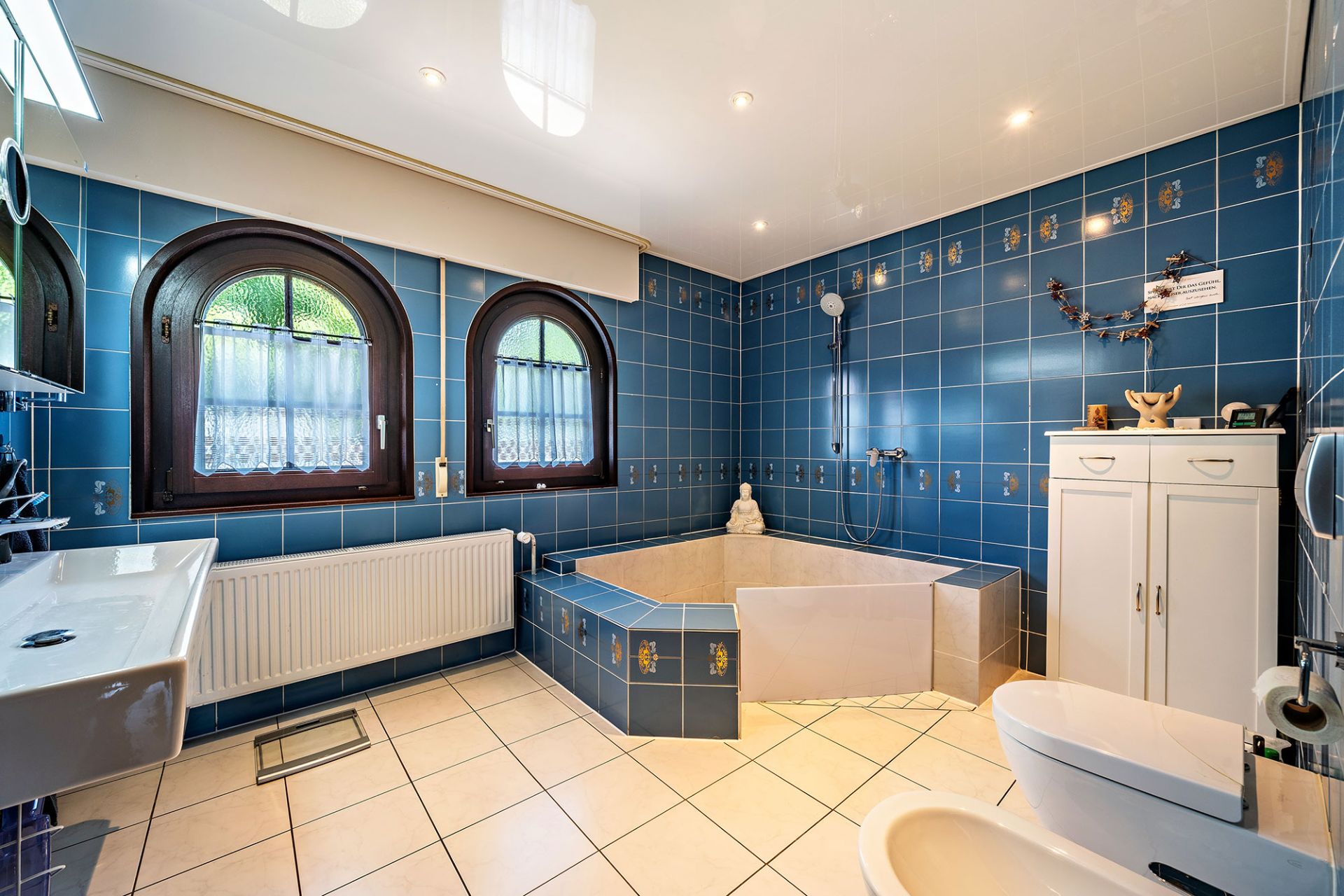 Modernes Badezimmer mit blauen Fliesen, Eckbadewanne, zwei Fenstern und weißen Sanitäranlagen.