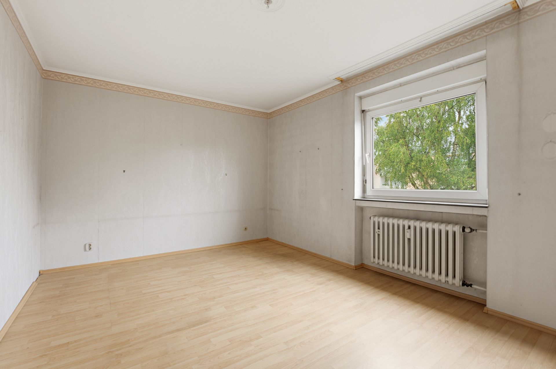 Leerer Raum mit hellem Laminatboden, großem Fenster und Heizkörper an der Wand.