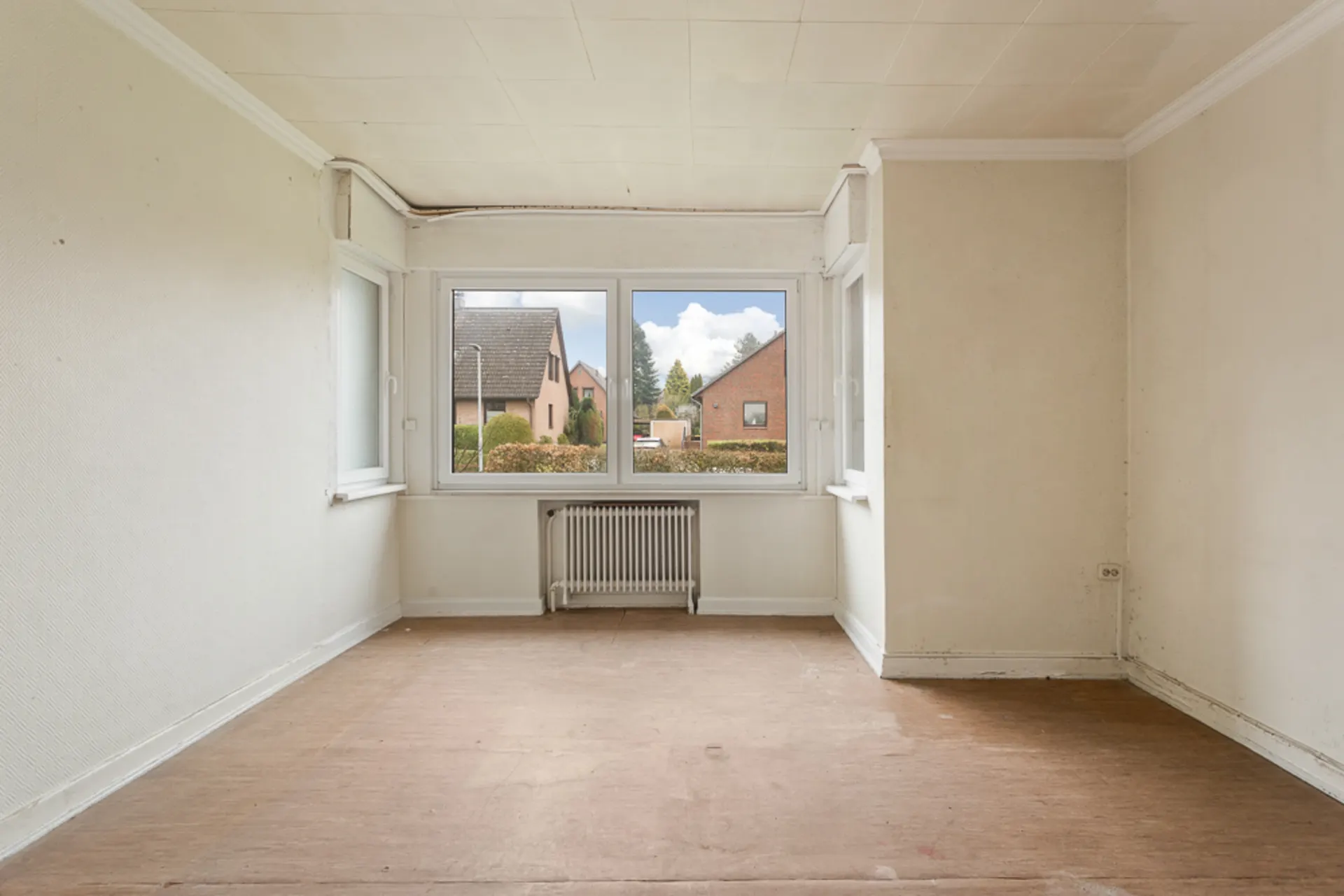 Leerer Raum mit großem Fenster, Blick auf Nachbarhäuser, unter dem Fenster steht ein Heizkörper.