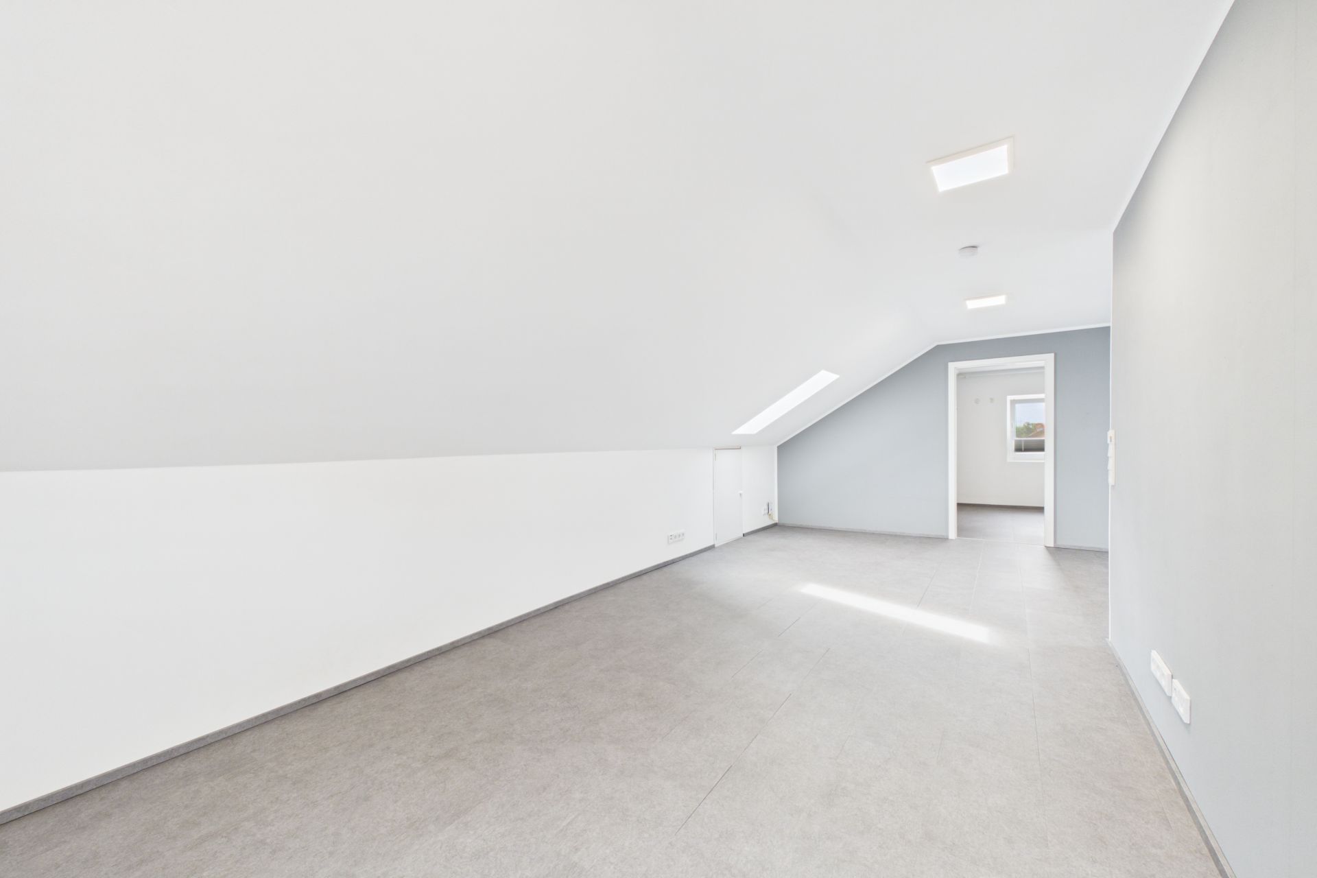 Heller, leerer Dachgeschossraum mit schräger Decke, weißen Wänden und grauem Boden.