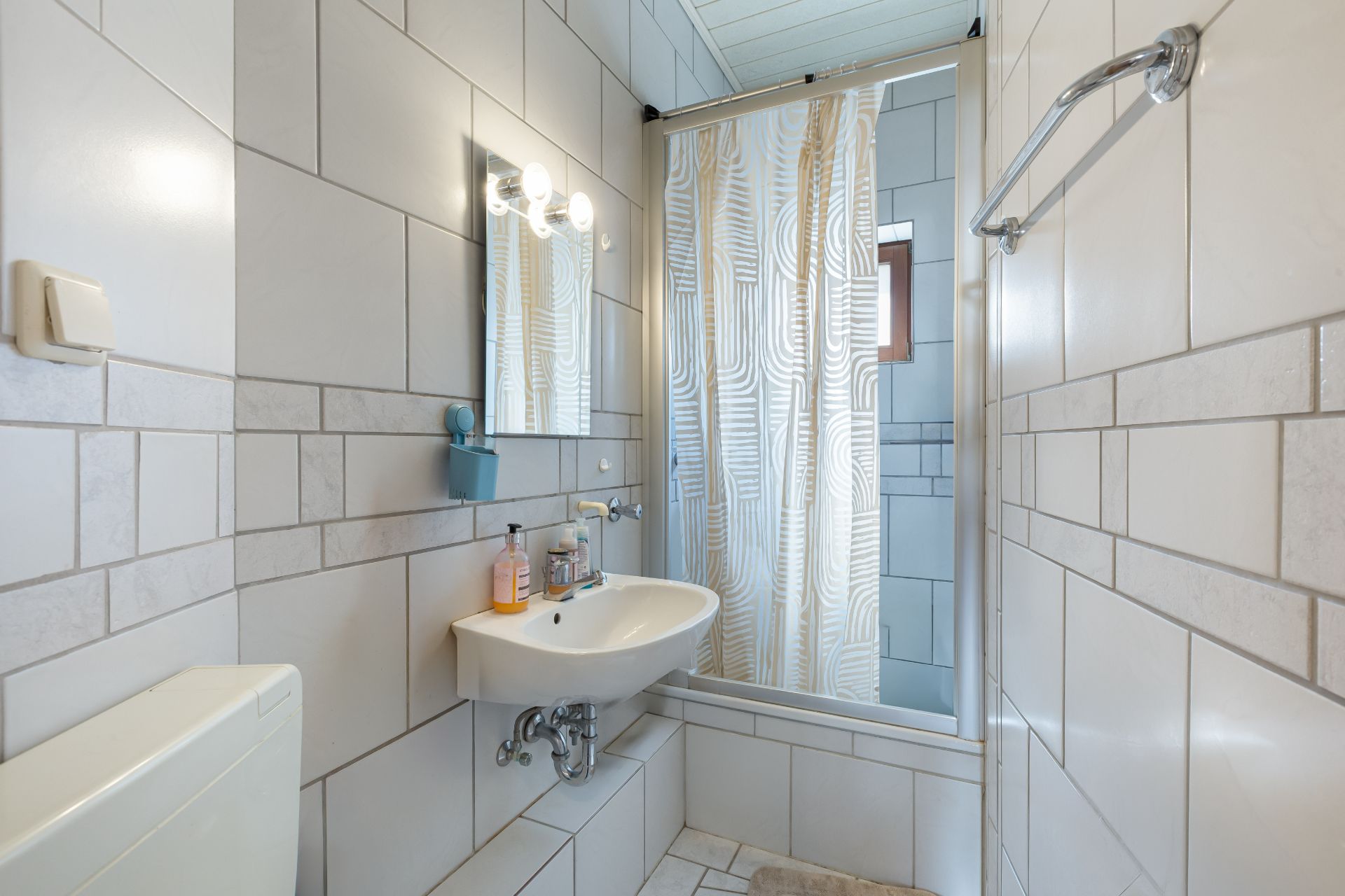 Kleines Badezimmer mit Waschbecken, Spiegel, Dusche mit Duschvorhang und weißen Fliesen.