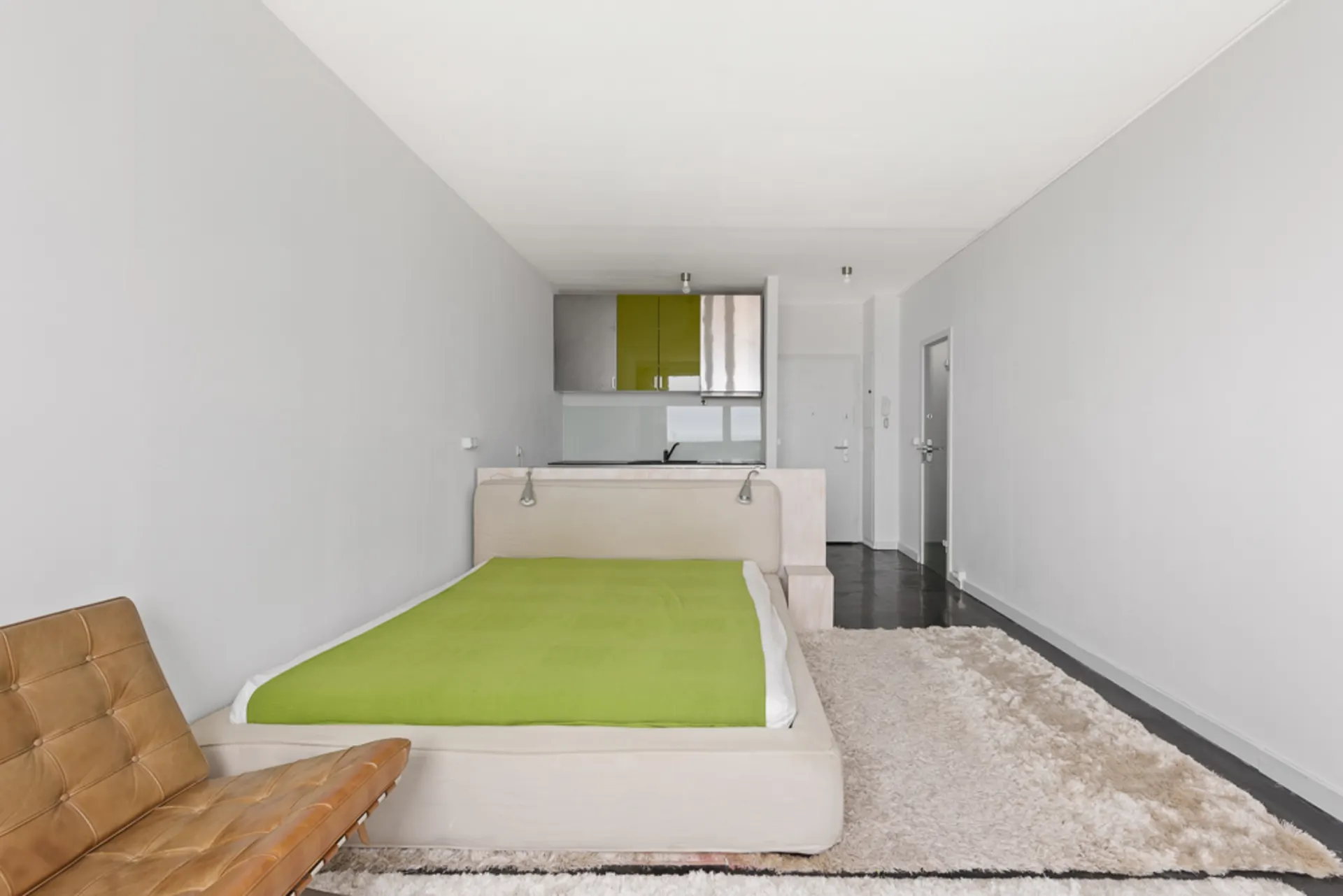 Modernes Schlafzimmer mit grünem Bett, braunem Sessel, Teppich und offener Küche im Hintergrund.