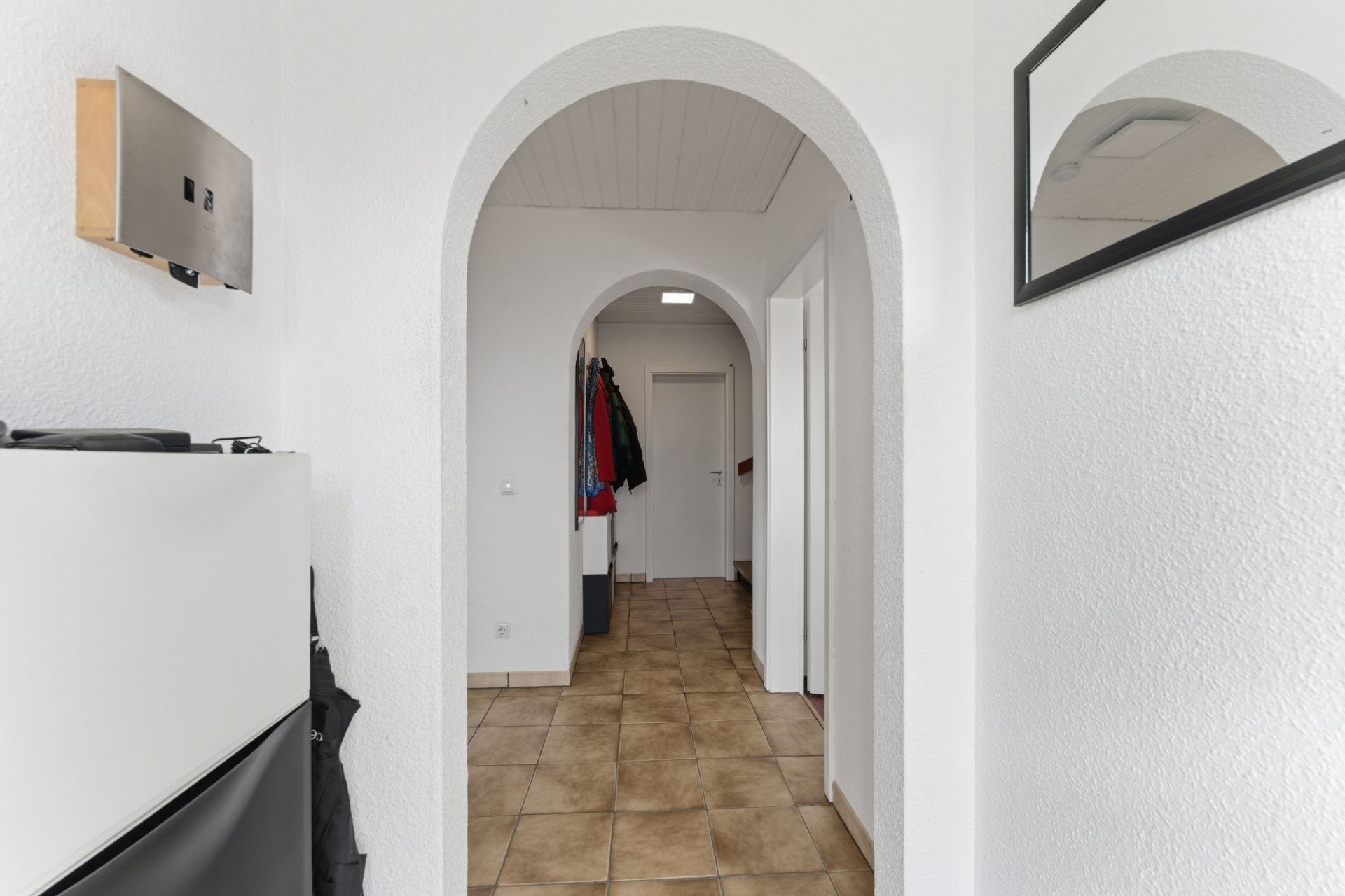 Flur mit weißen Wänden, zwei Rundbögen, Fliesenboden und Garderobe mit Jacken.