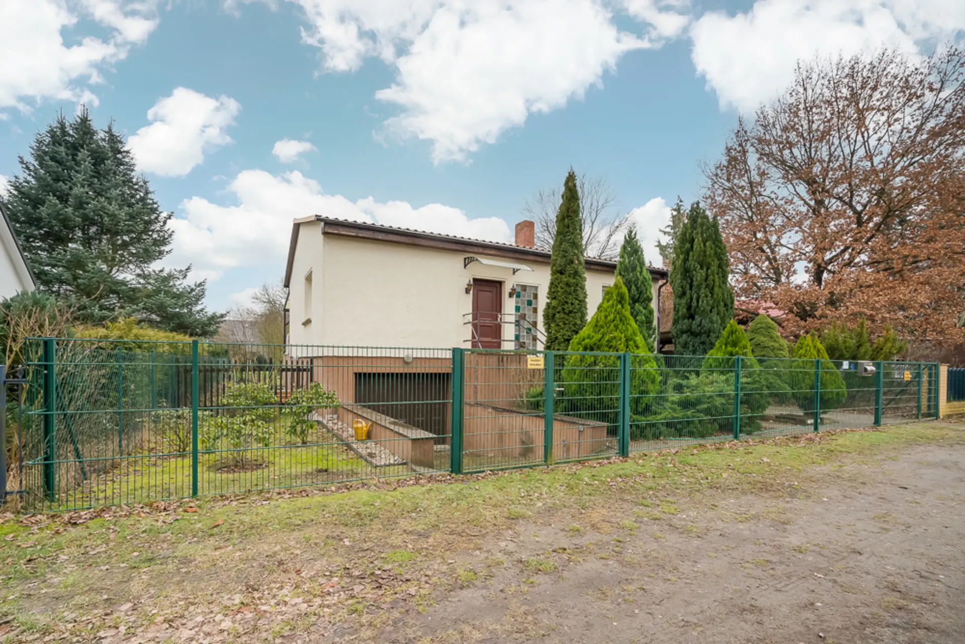 Einfamilienhaus mit Garten und grünem Metallzaun, umgeben von Bäumen und Sträuchern.