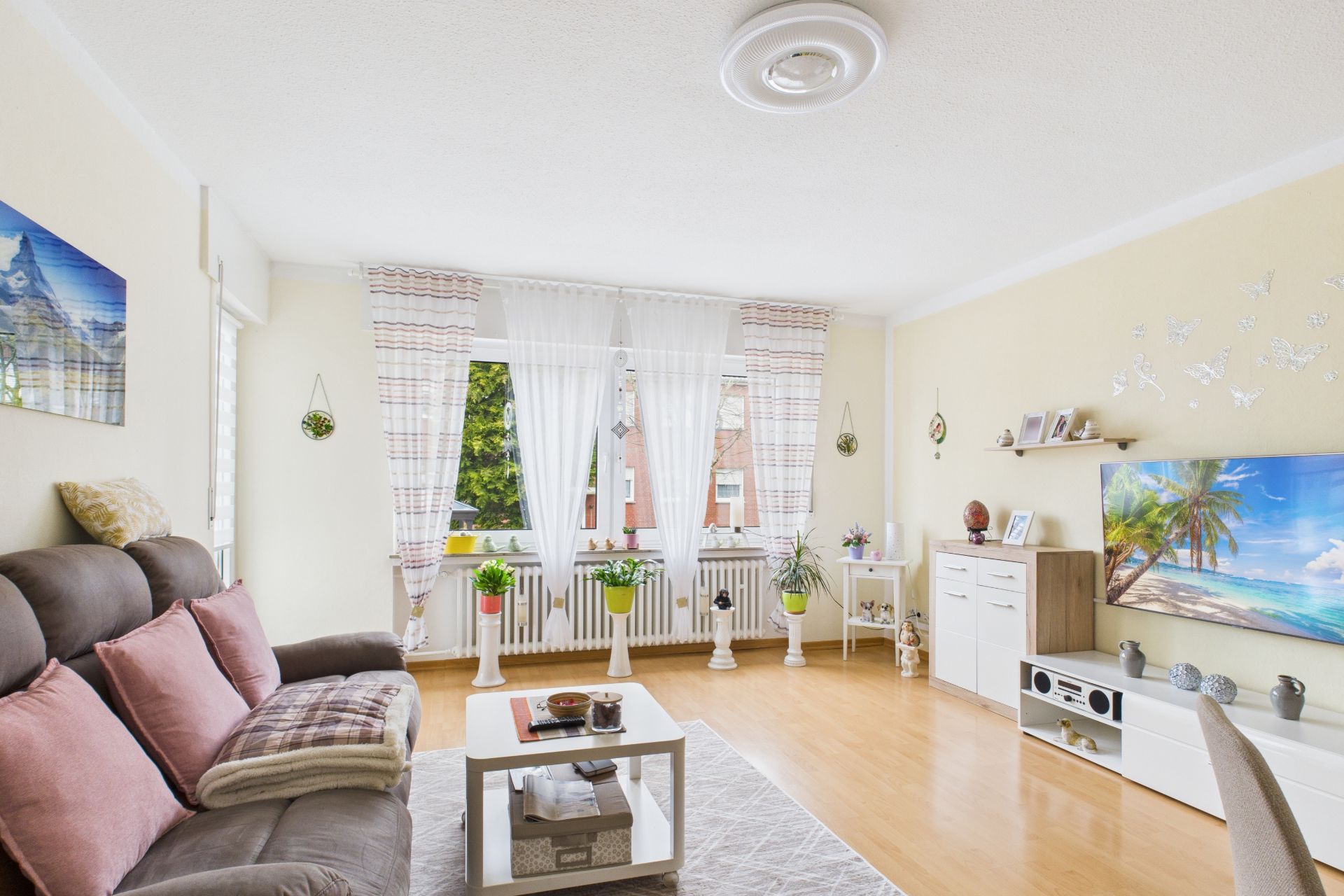 Helles Wohnzimmer mit Sofa, Couchtisch, TV, Pflanzen und großem Fenster mit Vorhängen.