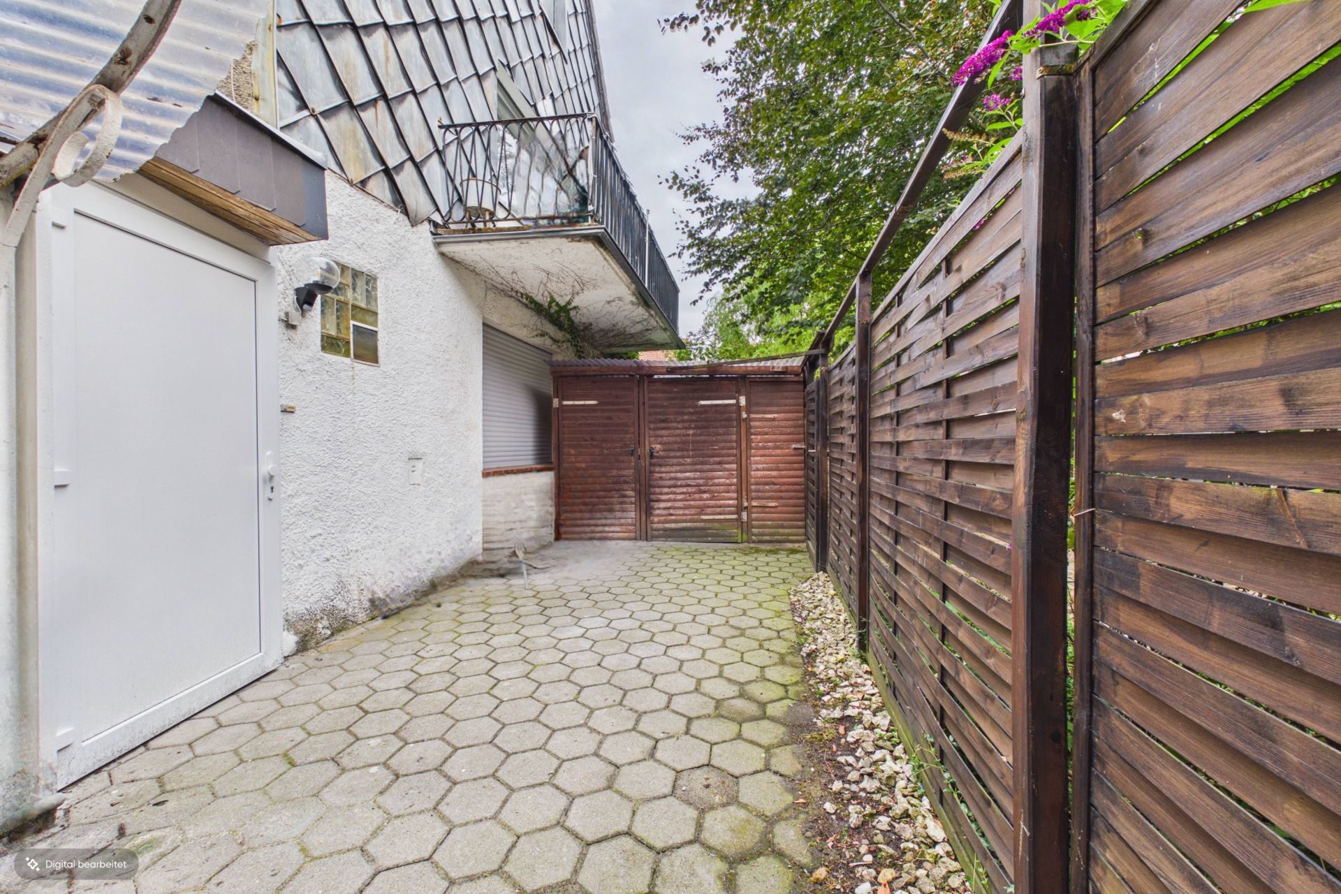 Gepflasterter Innenhof mit Holzzaun, Türen und Balkon über einer weißen Wand.