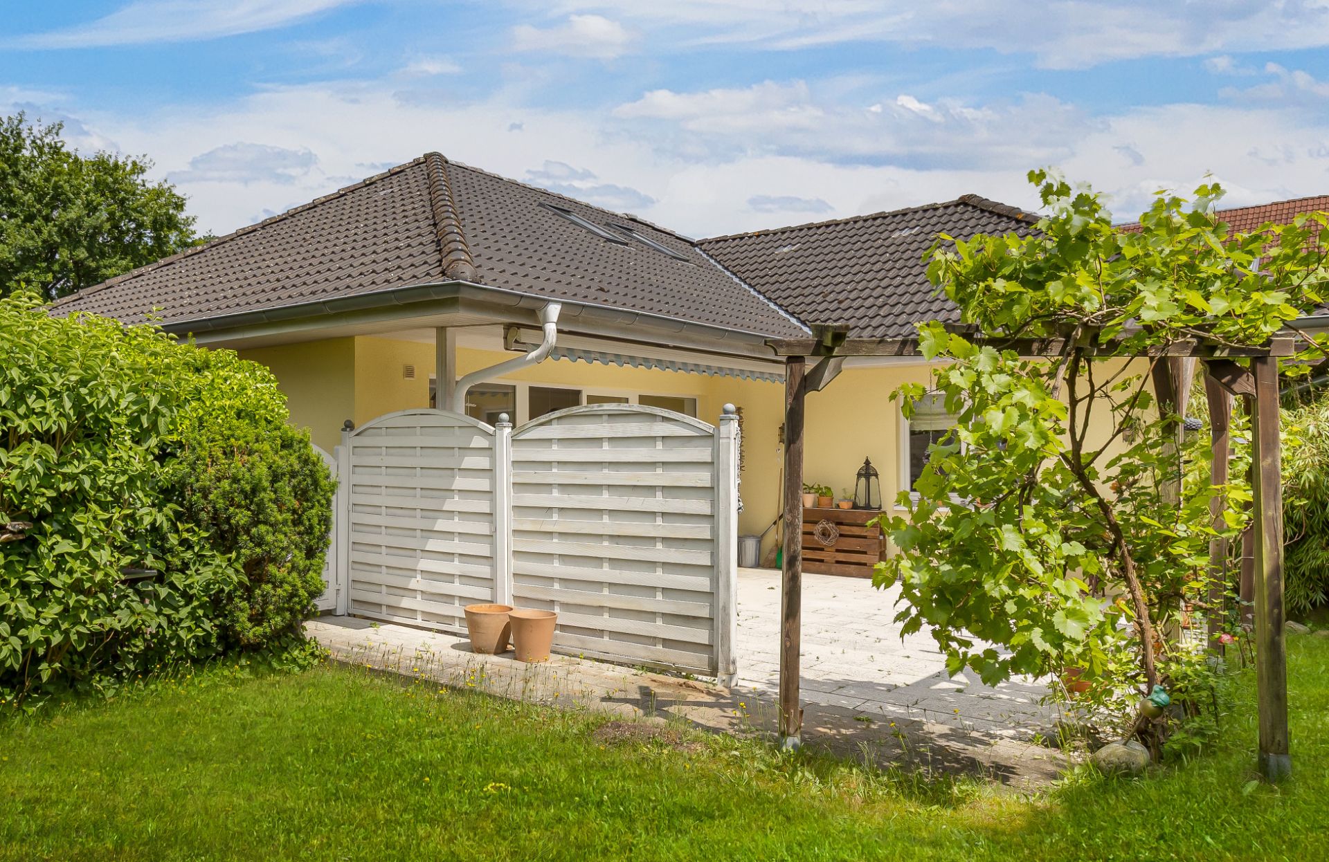 Garten mit Terrasse, Weinrebe, Holzzaun und gelbem Haus mit dunklem Dach im Hintergrund.