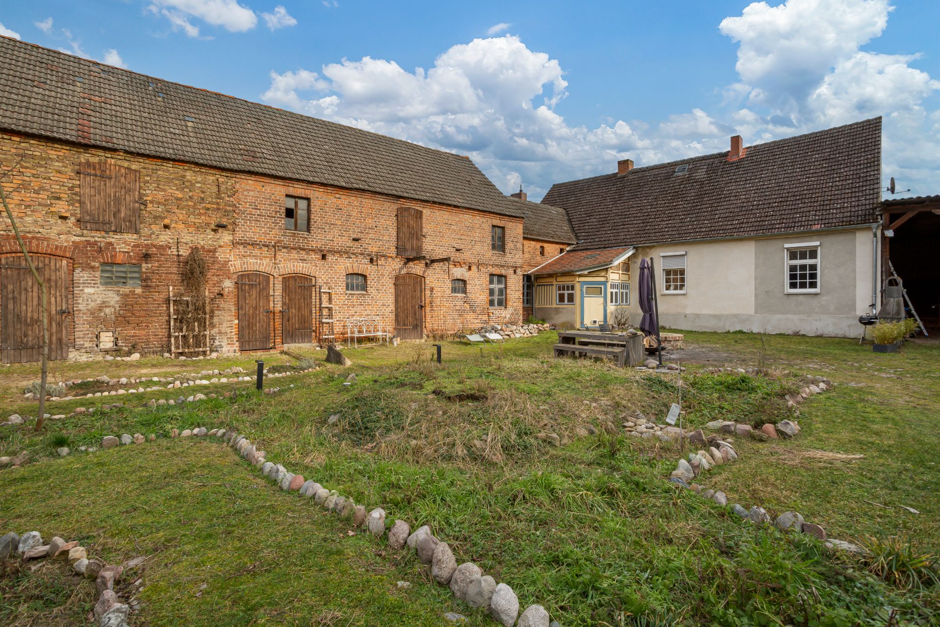 Großer Garten mit Rasen, steinernen Beetumrandungen und zwei Wohngebäuden im Hintergrund.