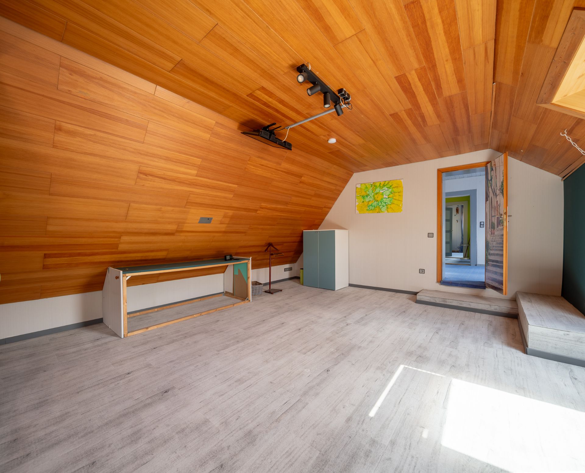 Helles Dachzimmer mit schräger Holzdecke, Schreibtisch, Schrank und offenem Durchgang.