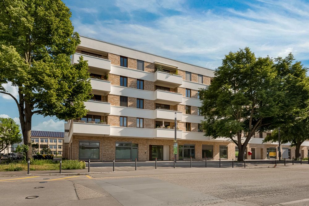 Modernes Wohngebäude mit vier Stockwerken und Balkonen, große Bäume davor.