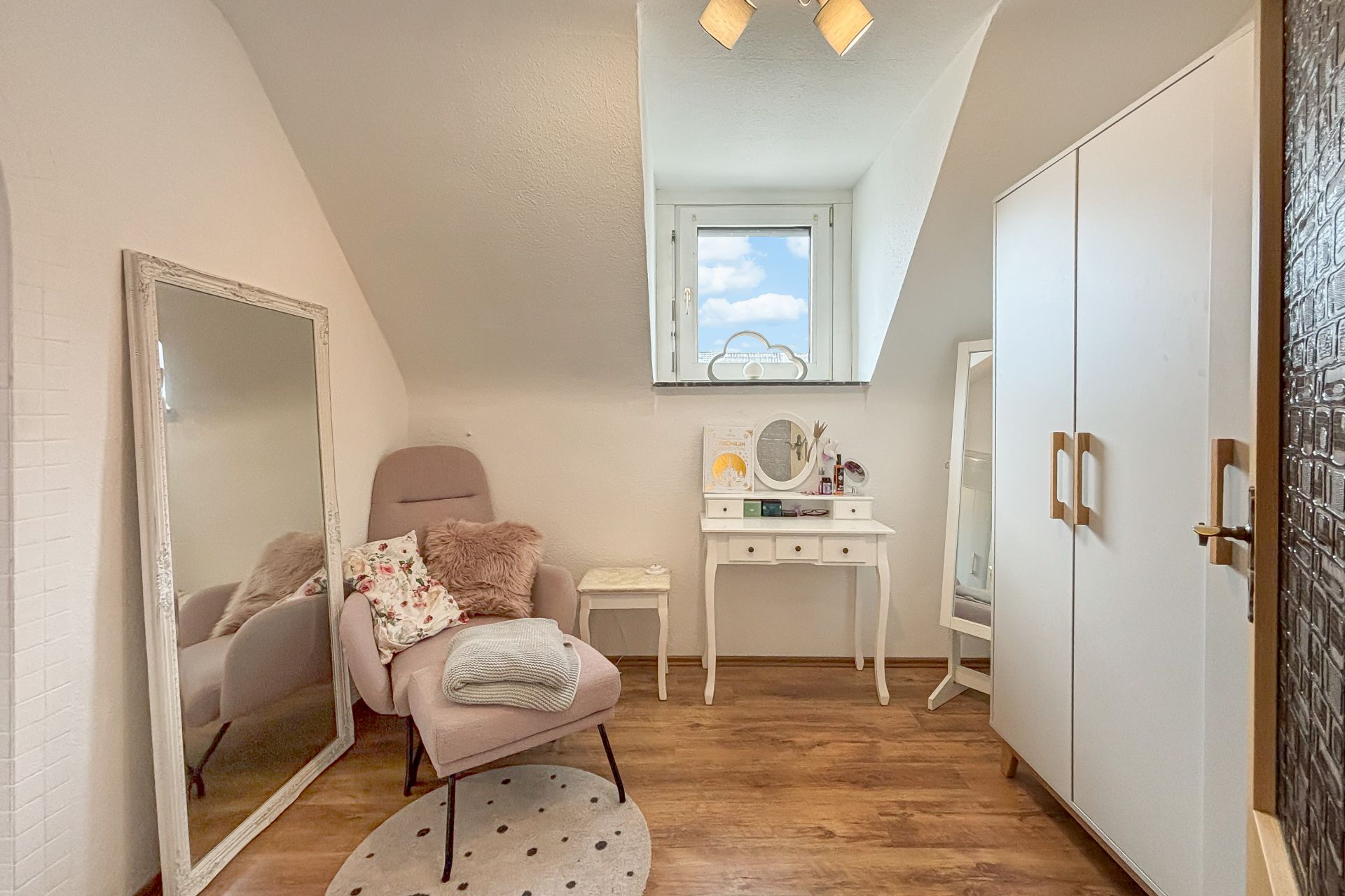 Helles Zimmer mit Sessel, Spiegel, Schminktisch und Kleiderschrank auf Holzfußboden.