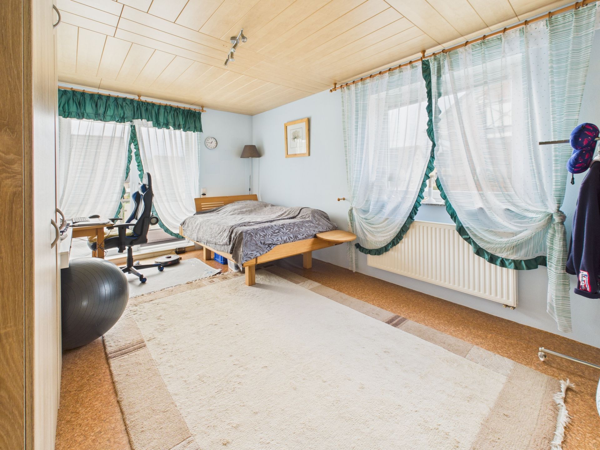 Helles Schlafzimmer mit Bett, Schreibtisch, Stuhl und großen Fenstern mit Vorhängen.