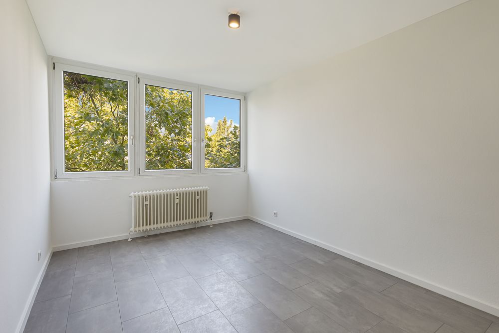 Heller leerer Raum mit drei großen Fenstern, grauem Fliesenboden und Heizkörper.