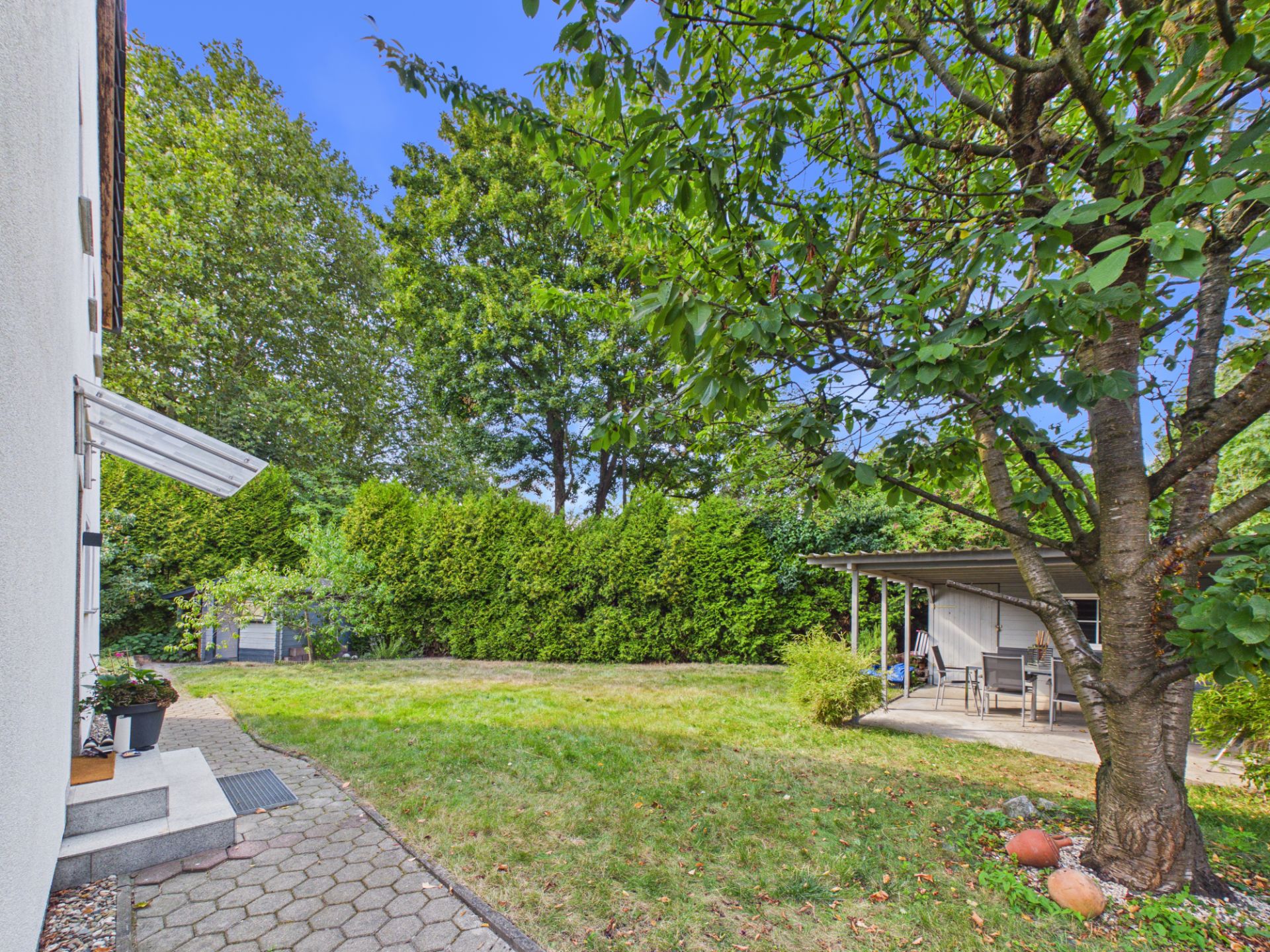 Garten mit Rasen, großem Baum, Terrasse mit Möbeln und angrenzendem Haus.