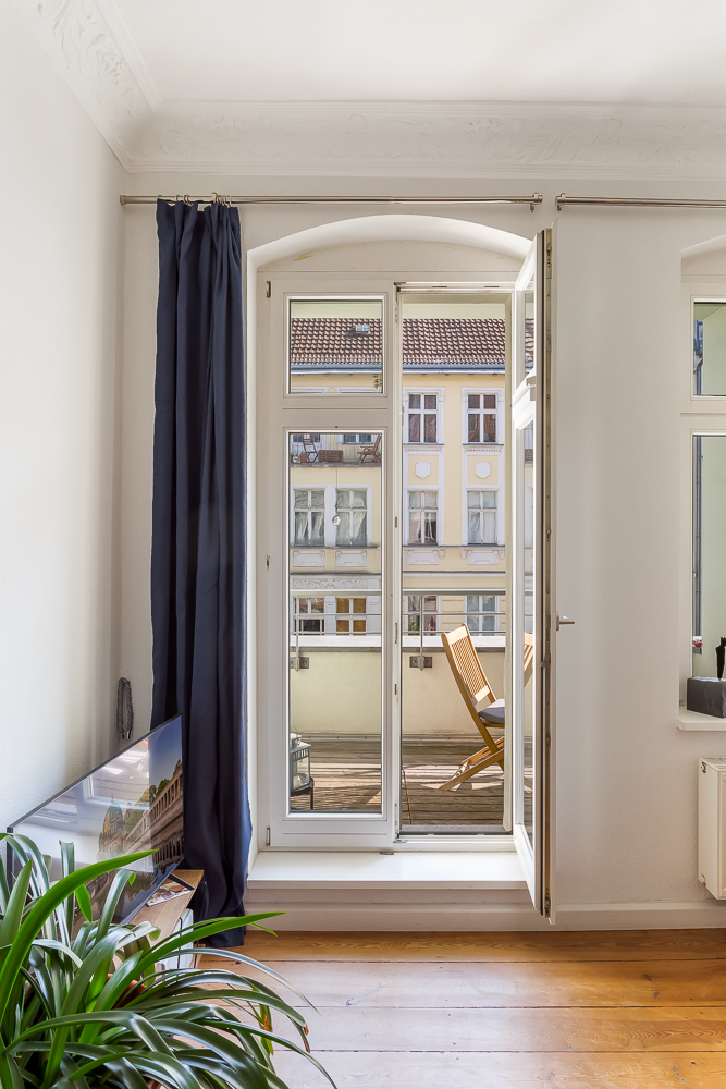 Geöffnete Balkontür mit Blick auf einen Holzbalkon und Nachbargebäude, Pflanze im Vordergrund.