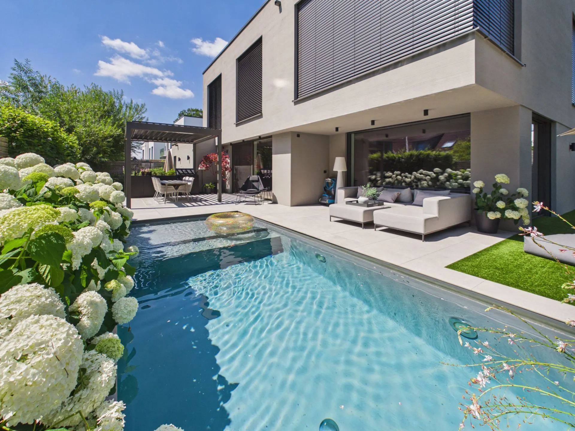 Moderner Garten mit Pool, Terrasse, Sitzgruppe und blühenden Hortensien vor einem Wohnhaus.