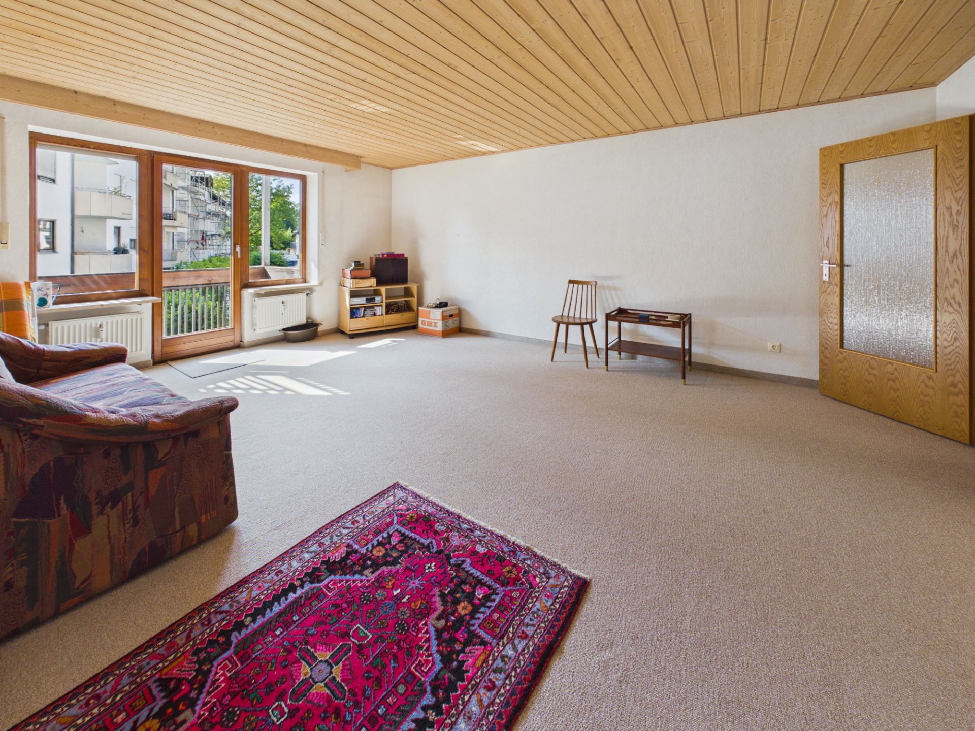 Helles Wohnzimmer mit Teppich, Sofa, Stuhl, Tisch und großer Fensterfront mit Tür zum Balkon.