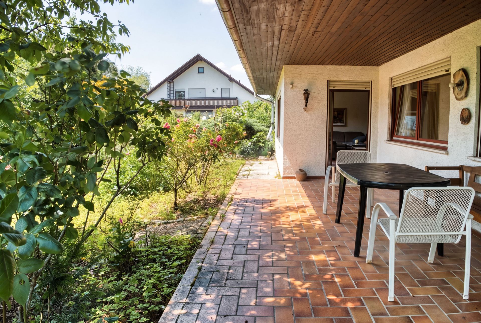Überdachte Terrasse mit Tisch und Stühlen, daneben Garten mit Sträuchern und Blumen.