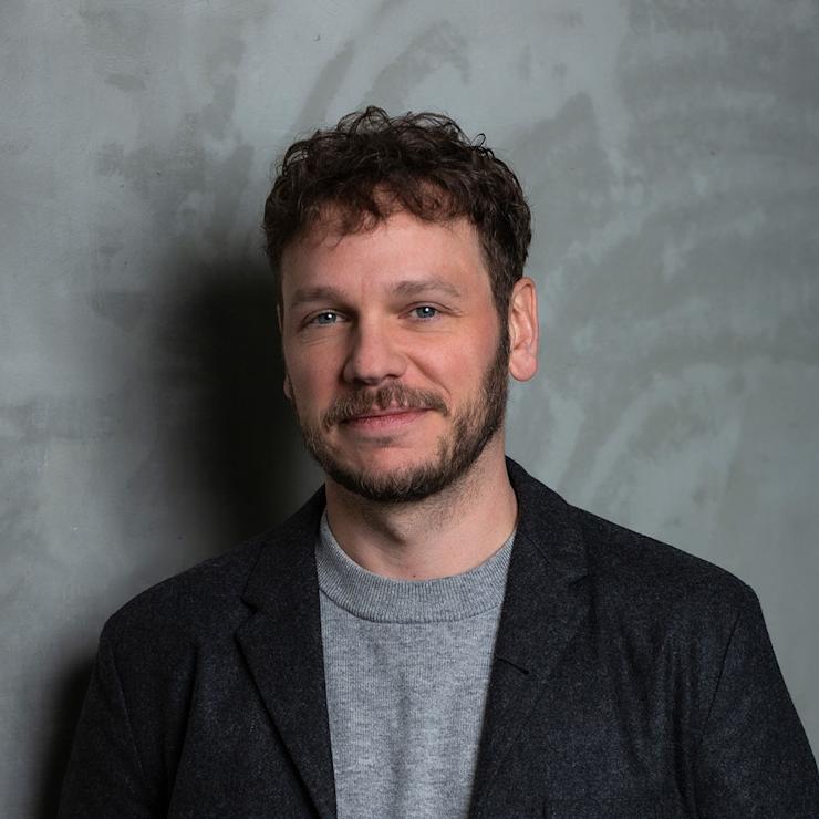 Ein Mann mit lockigen Haaren steht vor einer grauen Wand und trägt einen dunklen Blazer und ein graues Shirt.