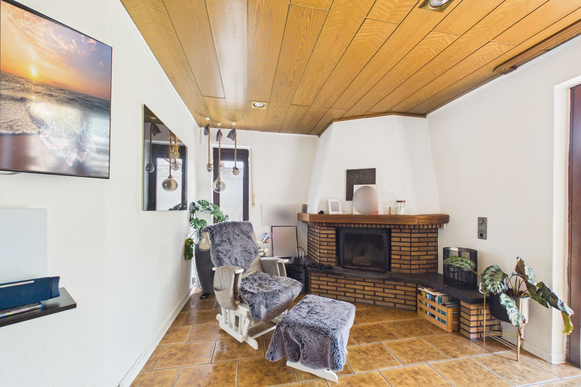 Wohnzimmer mit Kamin, Sessel, Pflanzen und Wandbildern, Holzboden und Holzdecke.