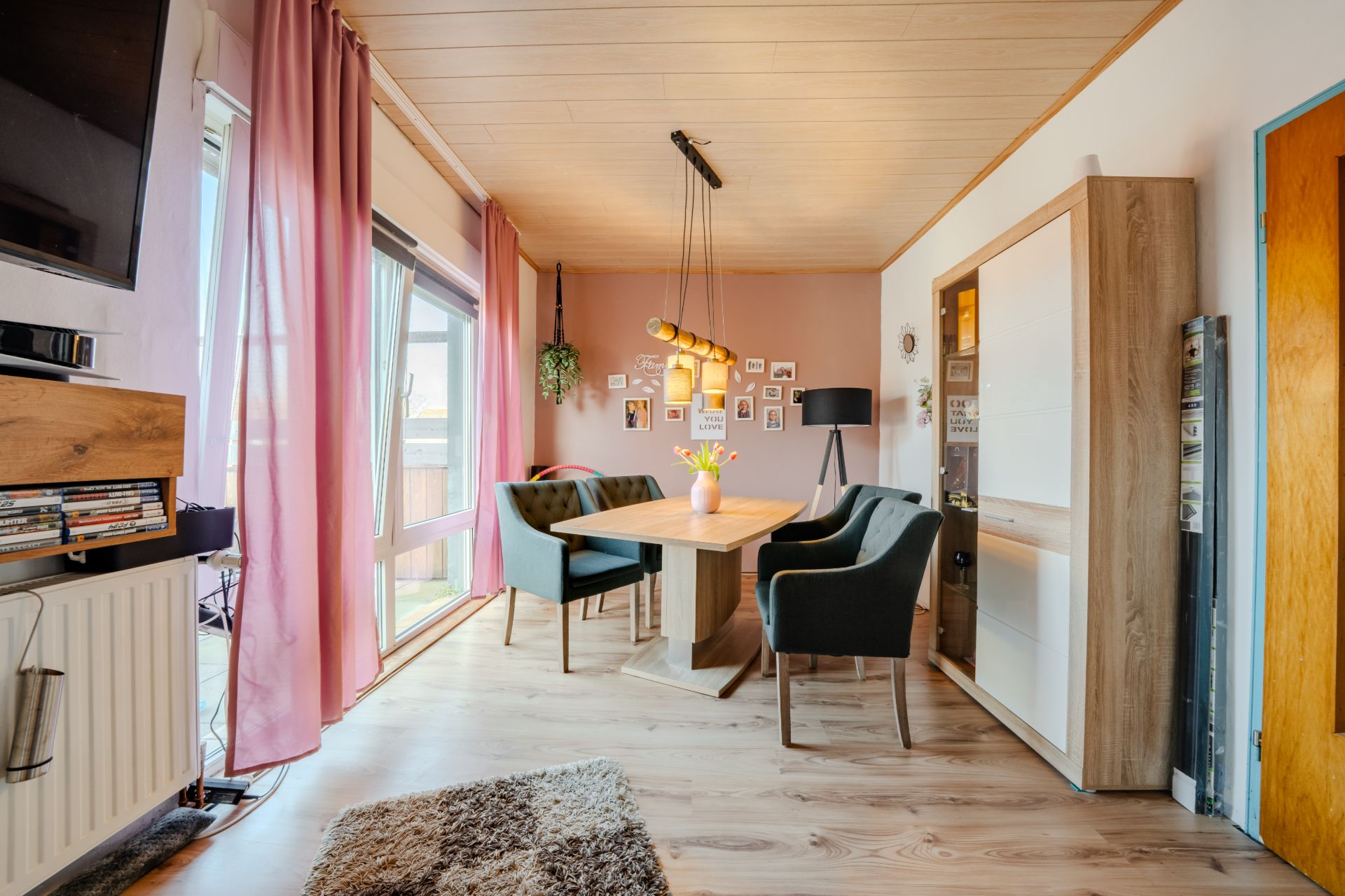Essbereich mit Holztisch, vier Stühlen, Regal, rosa Vorhängen und Wandbildern.