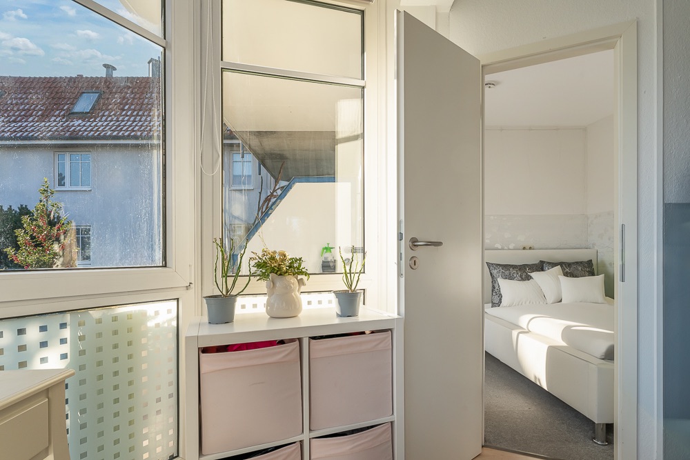 Helles Zimmer mit Pflanzen auf einer Kommode und Blick ins Schlafzimmer mit weißem Bett.