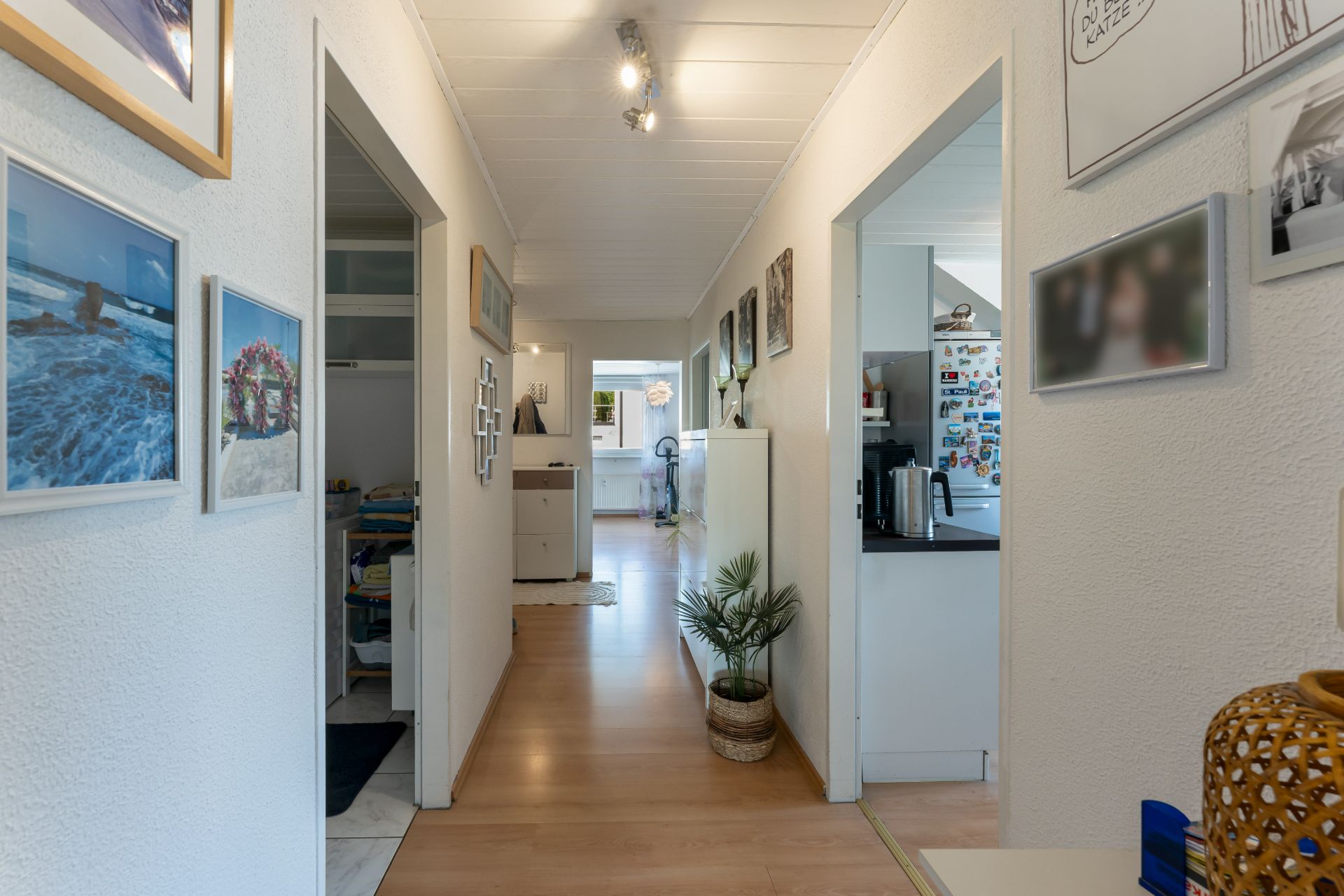 Langer Flur mit Holzfußboden, Pflanzen, Fotos an den Wänden und Zugang zu mehreren Zimmern.