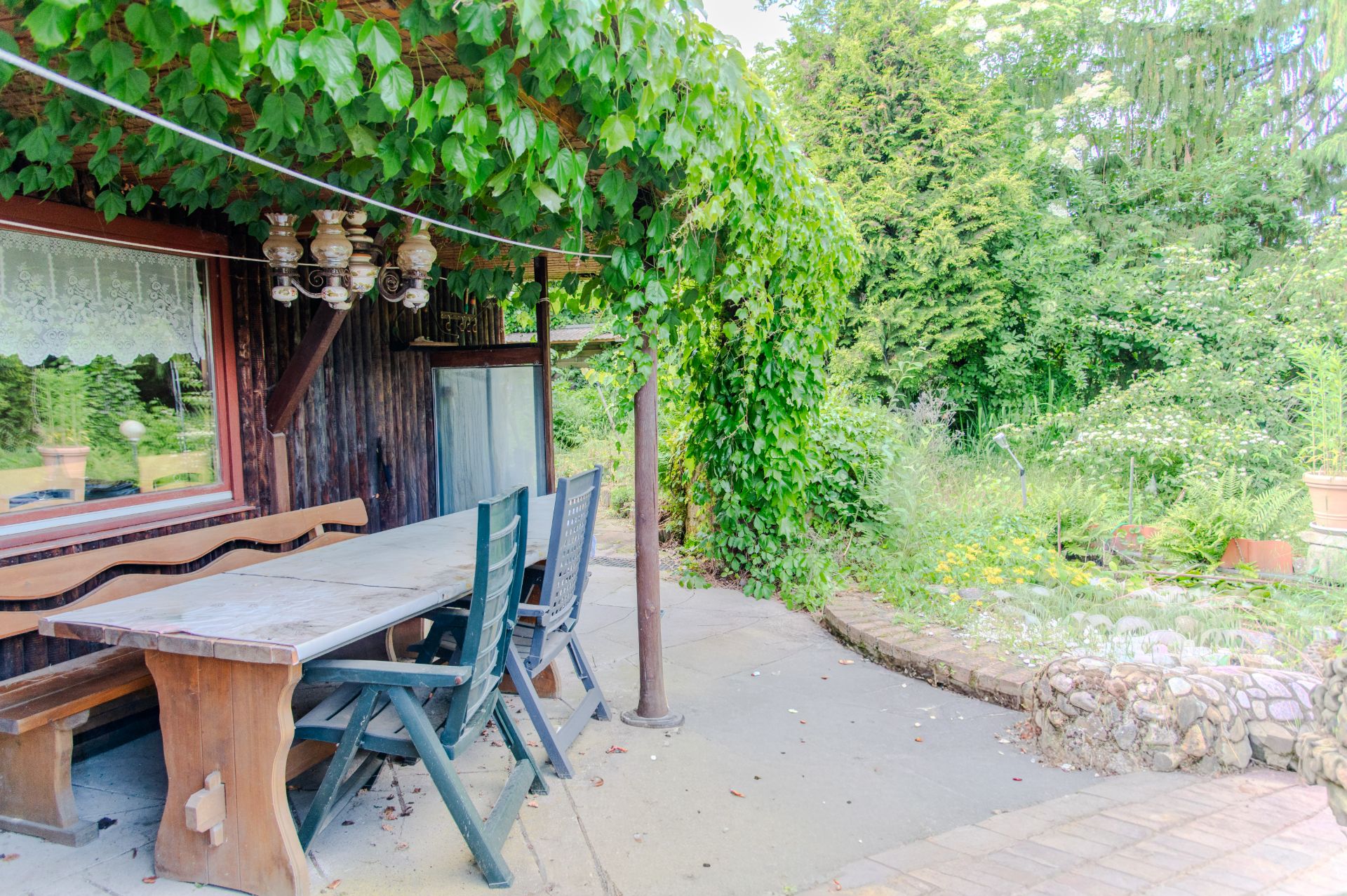 Überdachte Terrasse mit Holztisch, Bänken und Stühlen neben einem Garten mit viel Grün.