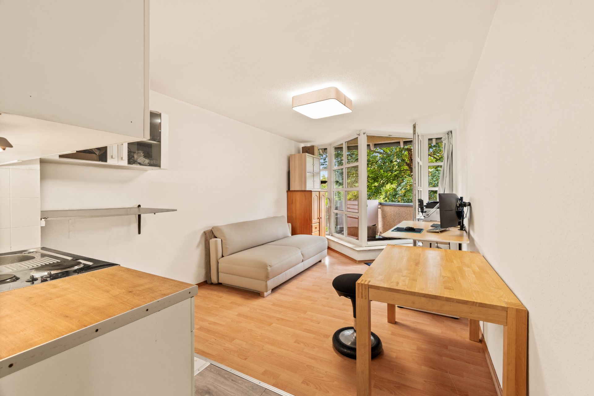 Helles Wohnstudio mit Sofa, Schreibtisch, kleiner Küche und Zugang zu einem Balkon.