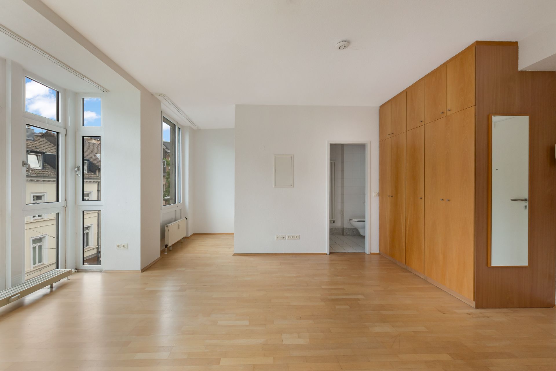 Helles Zimmer mit großen Fenstern, Holzfußboden, Einbauschrank und Zugang zu einem Badezimmer.