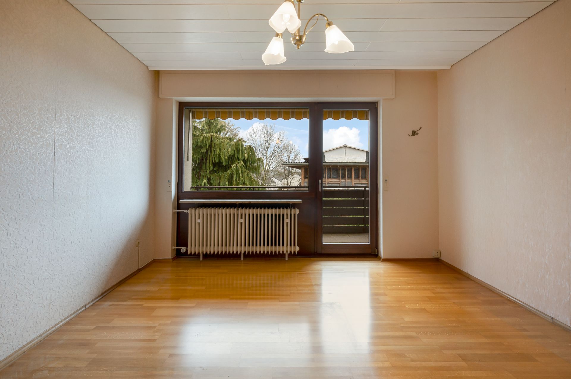 Leeres Wohnzimmer mit Holzfußboden, großem Fenster, Heizkörper und Deckenlampe.