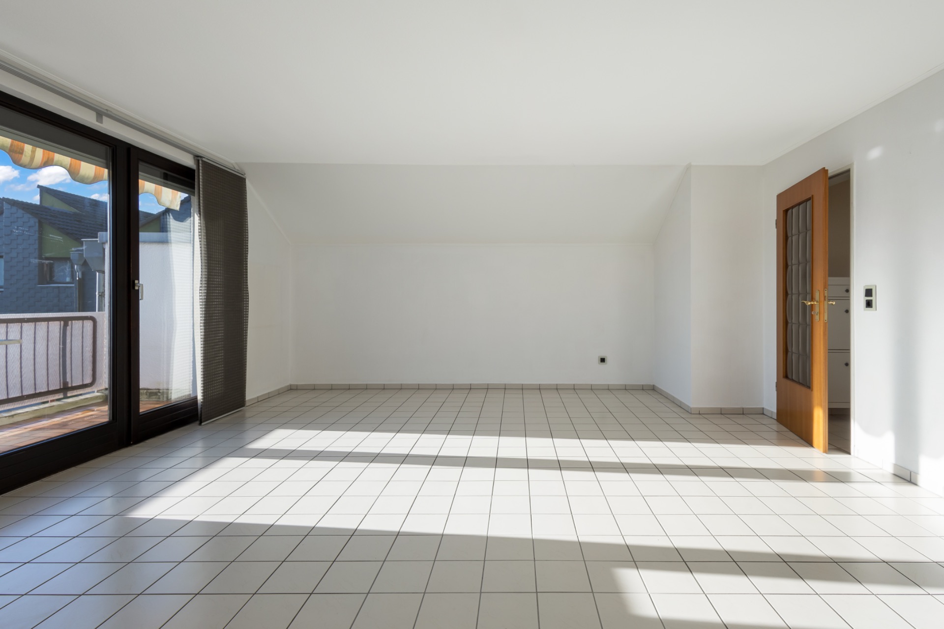 Helles, leeres Zimmer mit weißen Fliesen und Glastür zu einem Balkon.