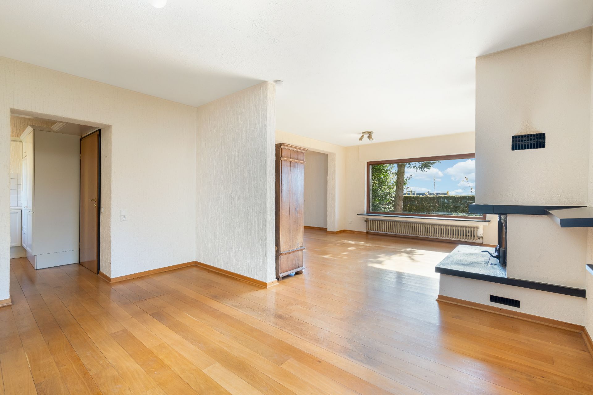 Helles Wohnzimmer mit Holzboden, Kamin, großem Fenster und Zugang zur Küche.