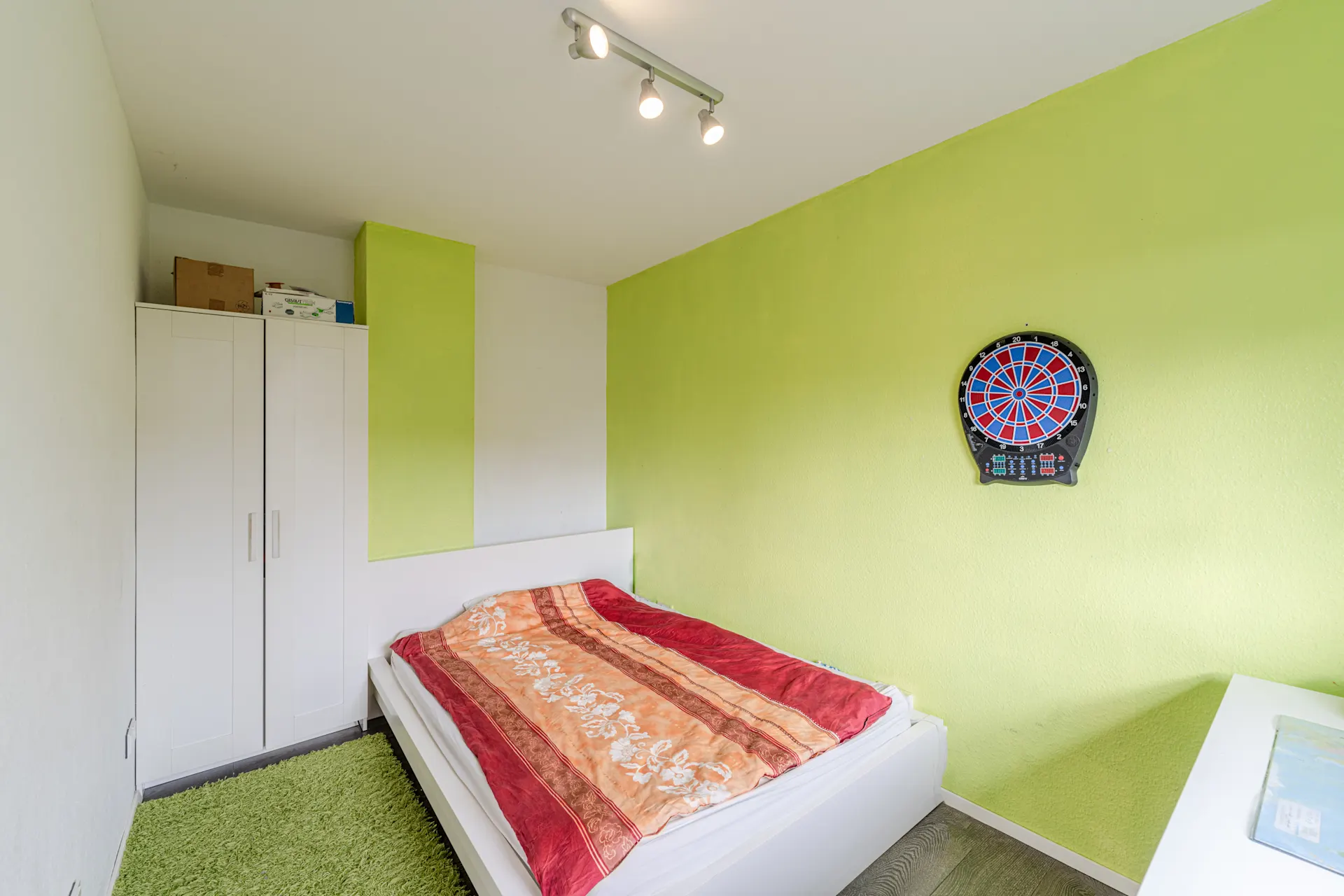 Schlafzimmer mit grün gestrichener Wand, Bett, weißem Schrank und einer Dartscheibe an der Wand.