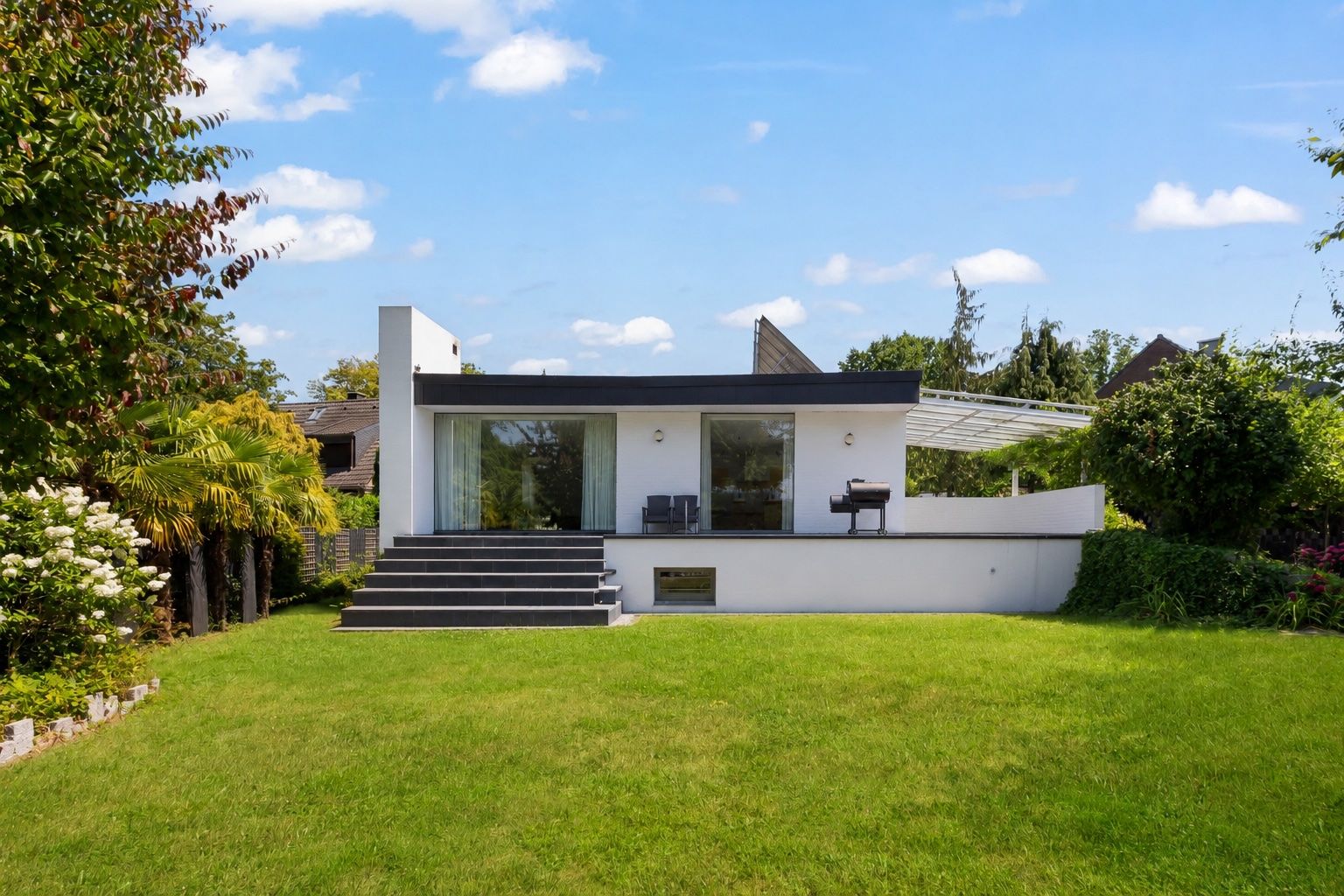 Modernes, einstöckiges Haus mit Terrasse und großem Garten, umgeben von Bäumen und Sträuchern.