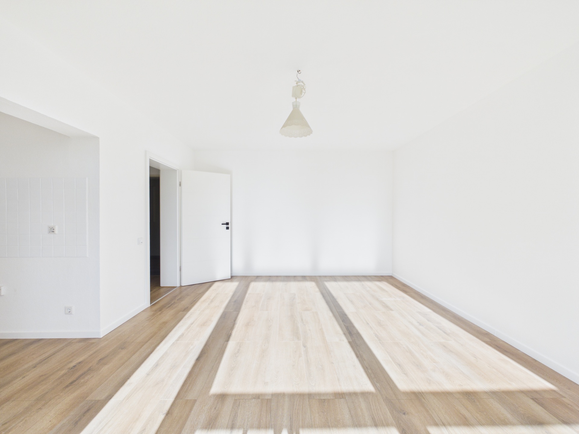 Helles, leeres Zimmer mit Holzfußboden, weißer Wand und offenem Türrahmen.