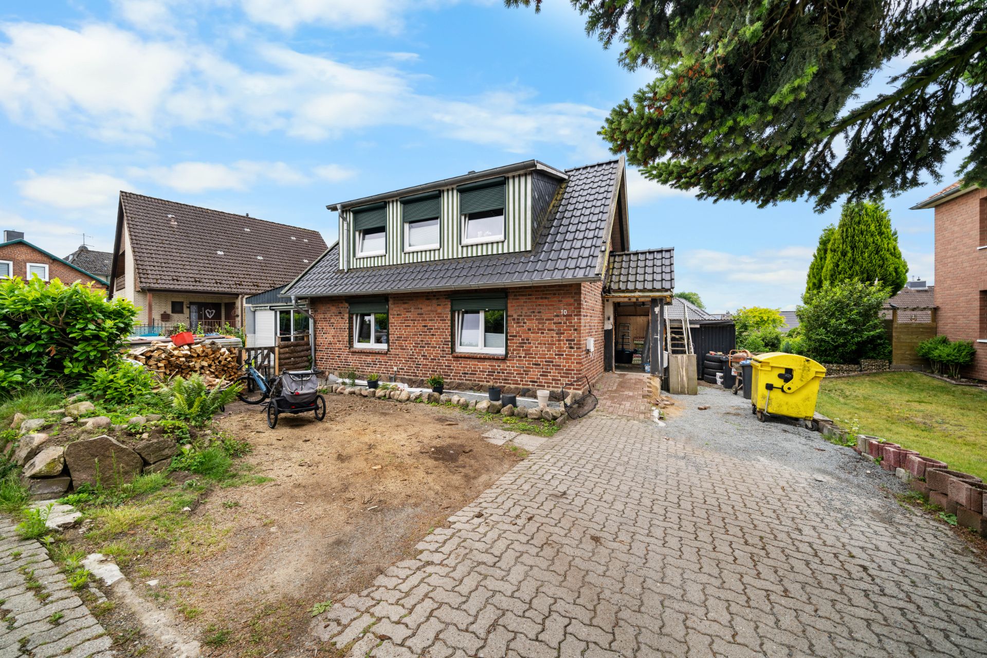 Backsteinhaus mit gepflasterter Einfahrt, Vorgarten und großen Fenstern, Gelbe Mülltonne rechts.