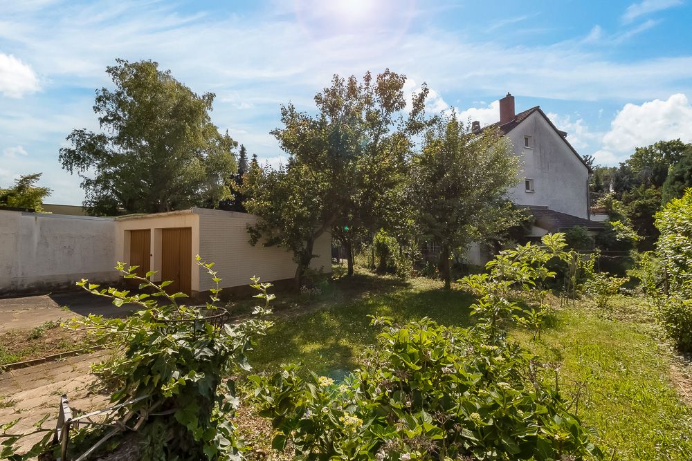 Grüner Garten mit Bäumen, Sträuchern und einer Garage neben einem Wohnhaus bei Sonnenschein.
