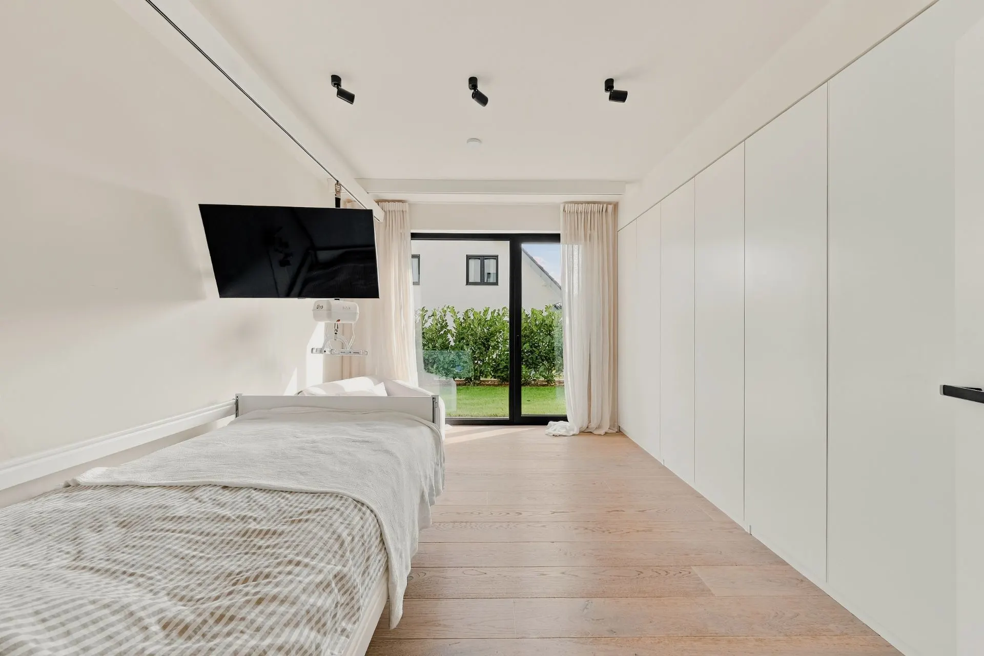 Helles Schlafzimmer mit Einzelbett, Fernseher, großen Einbauschränken und Fenster mit Blick in den Garten.