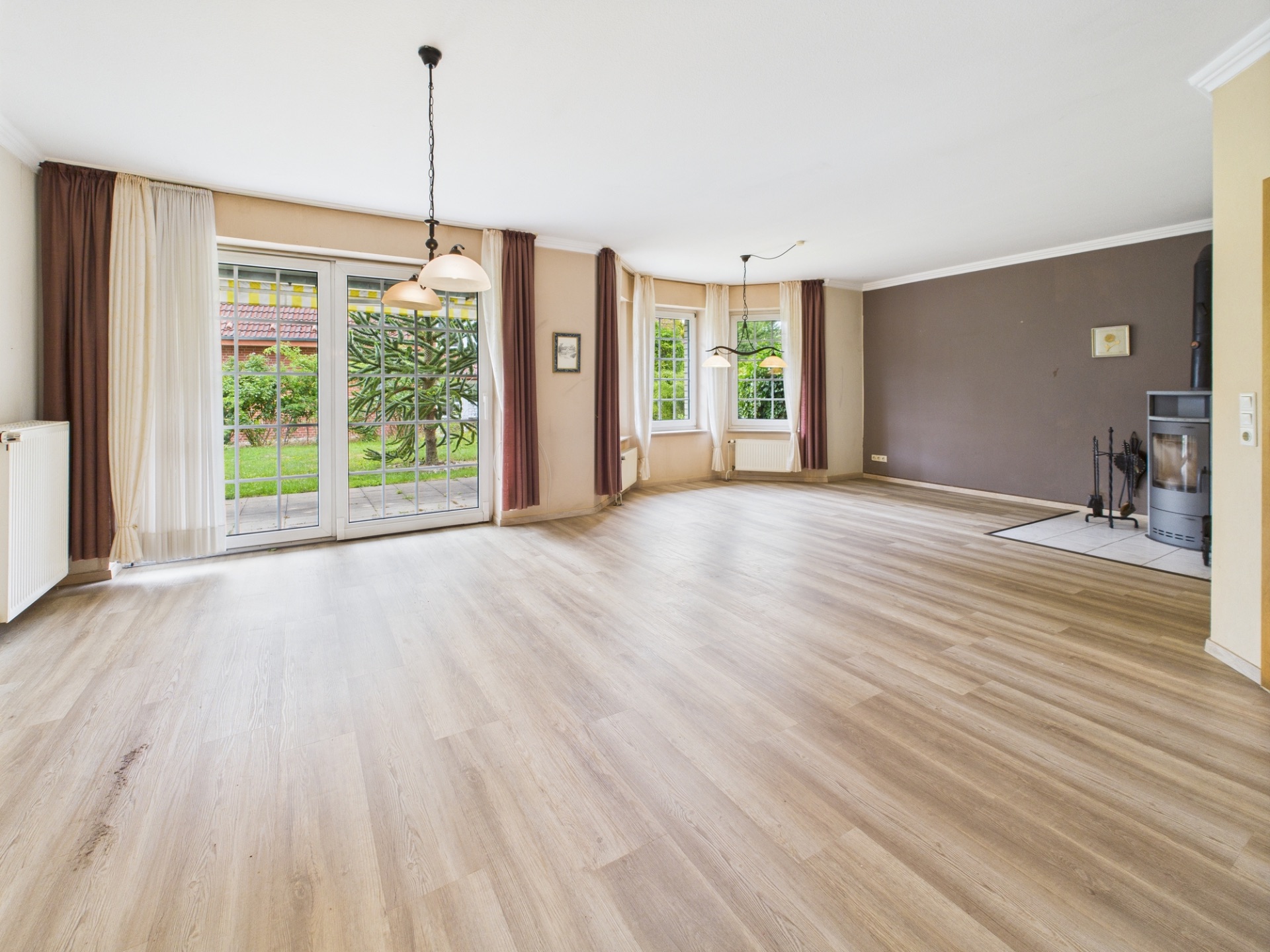 Helles, leeres Wohnzimmer mit großen Fenstern, Holzboden, Kamin und Blick in den Garten.